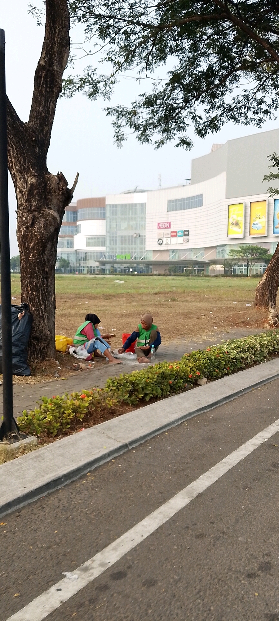 In Gunung Putri, Indonesia, two individuals, one wearing a pink and green vest and a black headscarf, the other a green vest and a beige headscarf, sit on a paved pathway. They appear to be sharing food or engaging in conversation. Behind them, a large tree trunk with dark bark is visible, with a black trash bag leaning against it. The ground is covered in dry grass and scattered debris. In the background, a modern, multi-story building with large glass windows and banners that include the "AEON" logo is prominent. The building is situated behind a grassy field and a line of green shrubs with red flowers separates the pathway from the road, which has a white line marking. The sky is overcast.