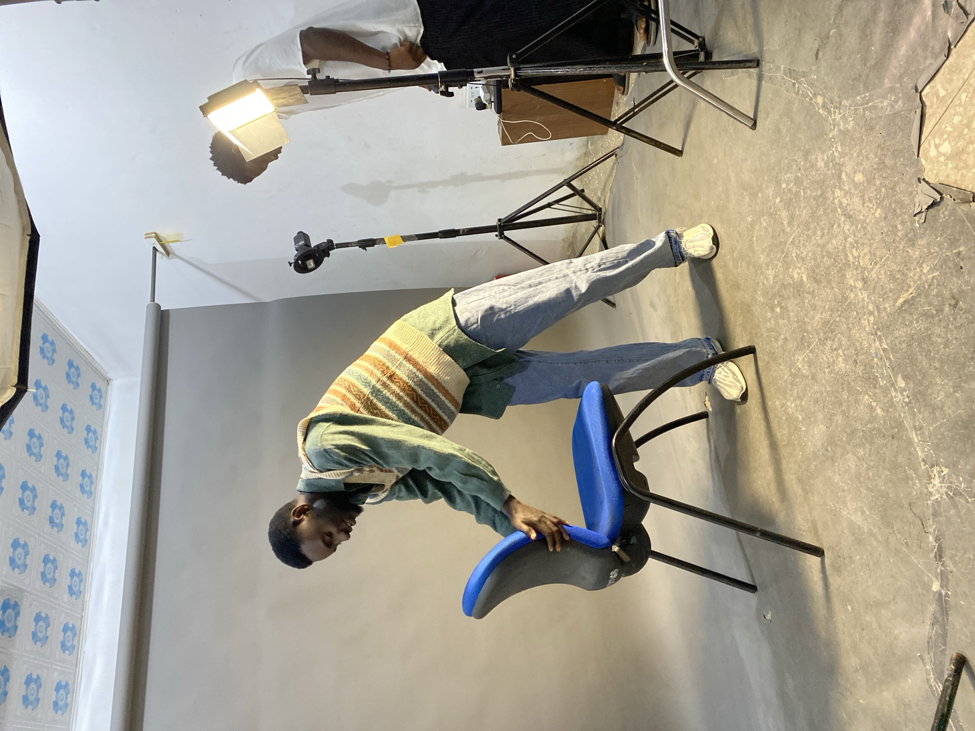 The image captures a behind-the-scenes moment of a photo shoot. A Black man is bent over, with one hand on a blue and gray office chair. He is wearing light blue jeans, a light green shirt, and a striped sweater vest. The scene appears to be set in a studio. Several lighting stands are visible in the background, including one directly overhead. The background consists of a grey wall and a patterned wall, with some light stands set up. The floor is concrete. The location is the city of Jos, Nigeria.