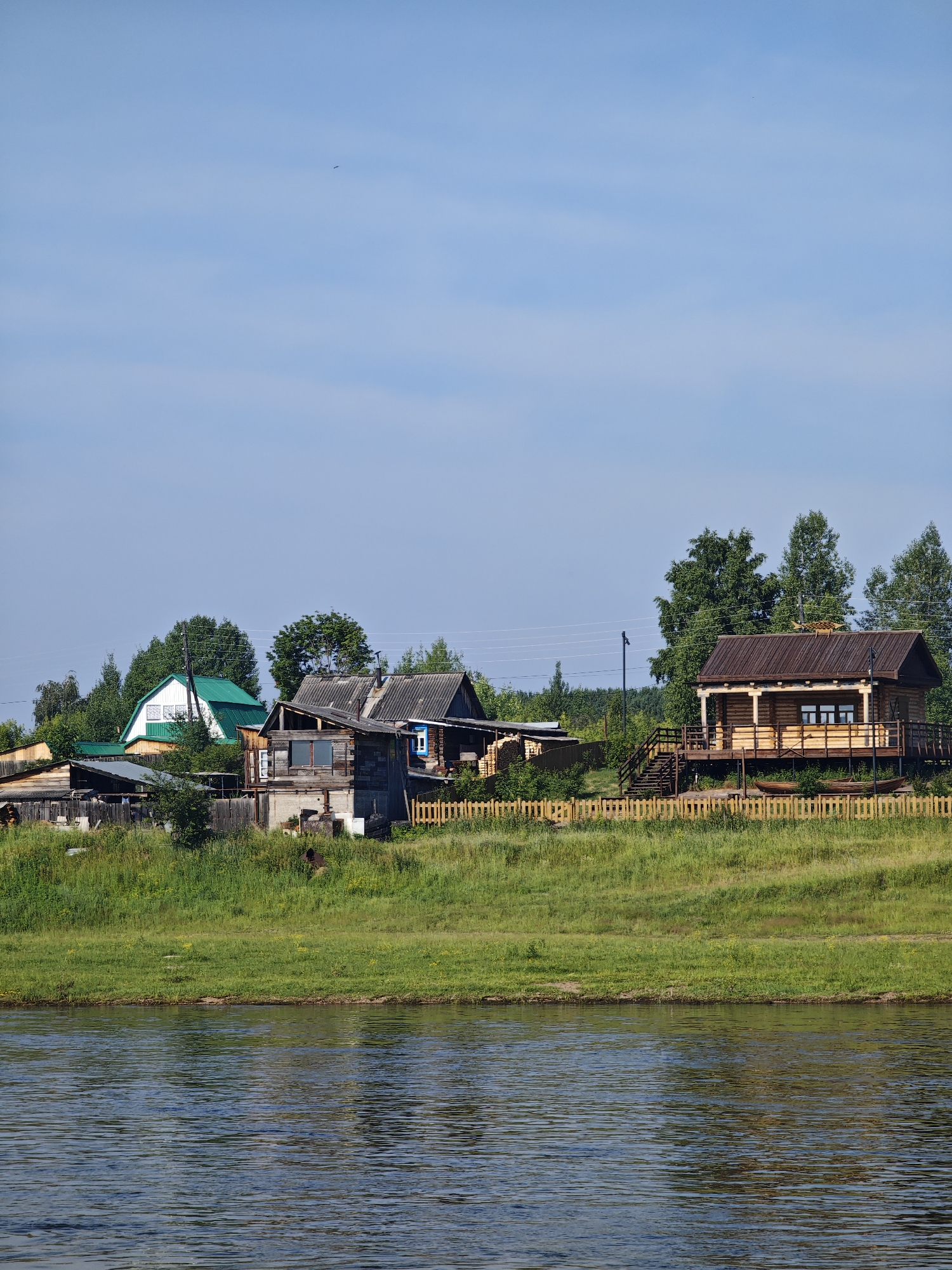 The image depicts a rural village scene in Galanino, Russia, on a clear day. In the foreground, a wide, calm river with a dark, reflective surface dominates the lower half of the frame. The riverbank is lined with lush green grass, which slopes upwards towards a cluster of rustic wooden houses and structures.
The village itself is situated on the elevated bank. Several wooden houses are visible, varying in architectural style and condition. One prominent structure on the right is a well-maintained log cabin with a wooden deck and stairs leading up to it. It has a dark-colored roof and several windows. To the left of the log cabin, there are older, more weathered wooden houses with corrugated roofs, some appearing to be in disrepair. One of these has a distinctive blue-framed window. Various outbuildings, fences, and stacks of lumber are scattered among the houses. A light-colored wooden fence runs along the crest of the bank, separating the village from the river.
The background is filled with dense green trees, forming a natural backdrop to the settlement. The sky is a clear, bright blue with a few wispy clouds and a distant, tiny dark speck that could be a bird. The overall atmosphere is one of peaceful rural life, with the river and the surrounding nature playing a significant role in the setting.