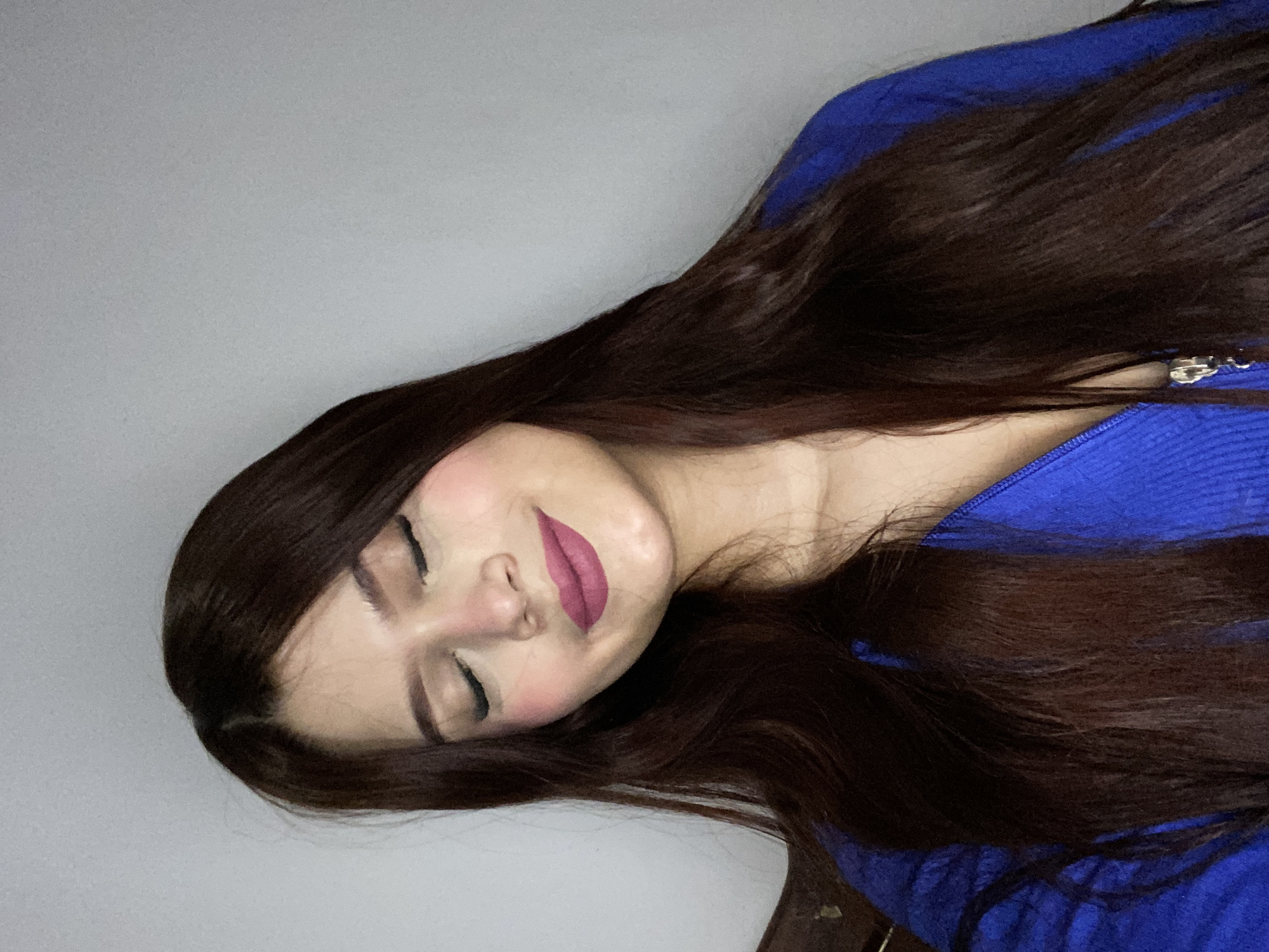 An indoor portrait features a young woman lying on her side, with her head resting on a surface and her body extending diagonally across the frame. Her eyes are closed, and she has a gentle, peaceful smile, suggesting a relaxed or sleeping state. Her long, dark brown hair, with subtle reddish highlights, cascades around her head and shoulders, covering part of her chest and the background.
She is wearing a vibrant royal blue, ribbed long-sleeved top or sweater, which appears to have a zipper at the front, visible near her shoulder. Her face is made up with dark eyeliner or eyeshadow, defined eyebrows, blush on her cheeks, and a prominent dark pink lipstick. The skin tone is fair.
The background is a plain, light gray wall or surface, largely out of focus, creating a clean and minimalist setting. The lighting is soft and even, highlighting her features without harsh shadows. There are no visible location cues within the image itself to suggest the provided context of Juan Griego, Venezuela. No text is visible in the image.