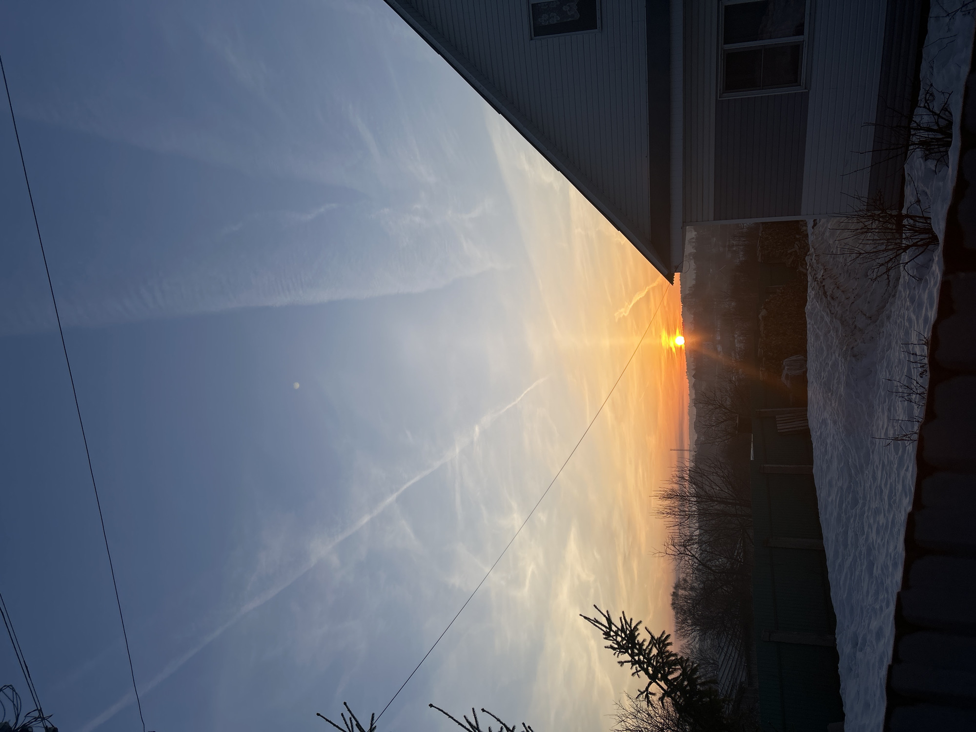 The image depicts a sunset in Фоки, Russia. A white building occupies the upper right corner of the frame, with a section of its wall visible. The sky transitions from blue to orange as the sun sets on the horizon. Wispy clouds streak across the sky. In the foreground, trees and a snow-covered fence can be seen. A wire stretches diagonally across the sky.