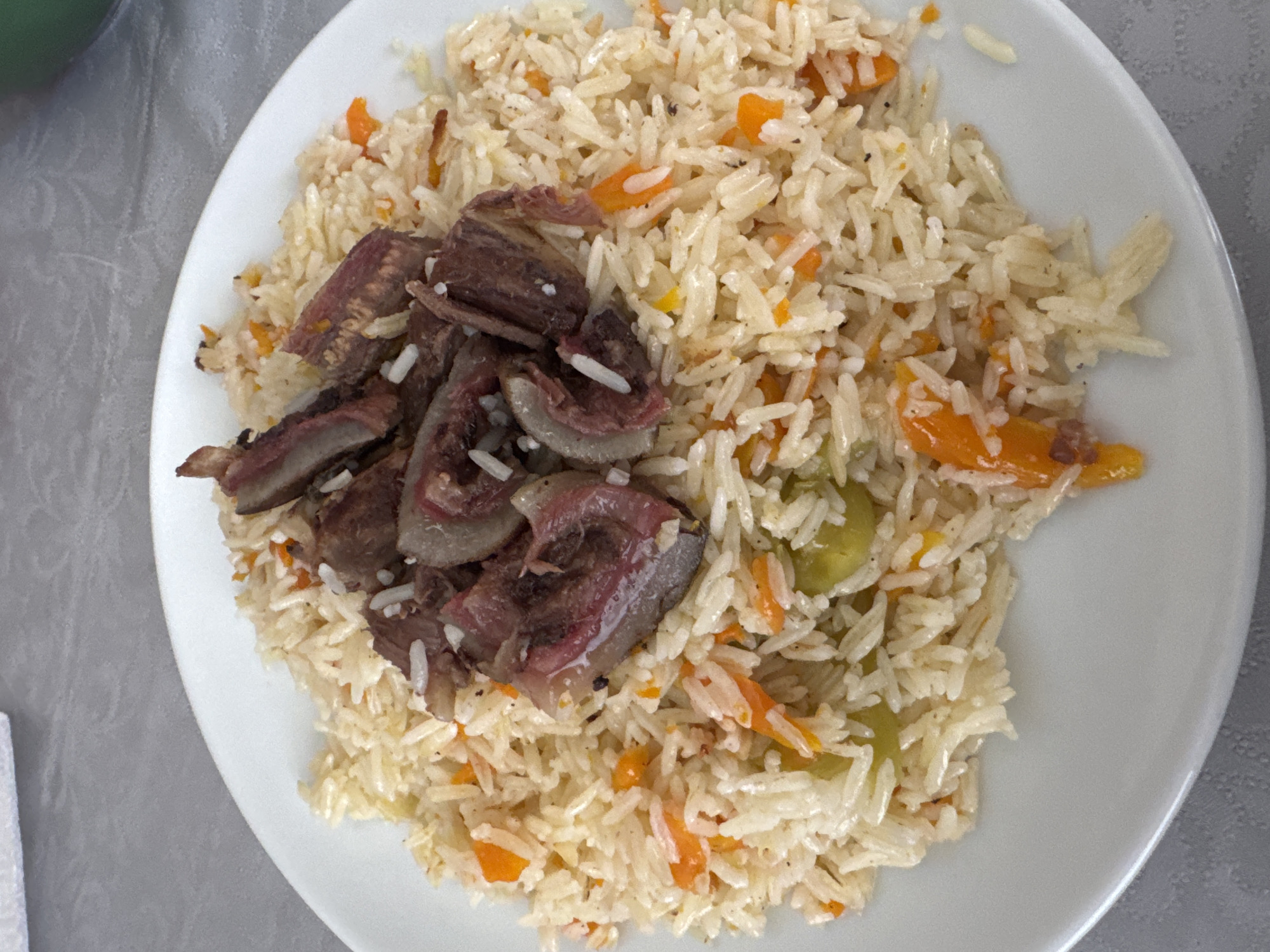 A close-up, overhead shot shows a white plate filled with pilaf topped with meat. The pilaf is composed of long-grain rice, diced orange carrots, and what appear to be pieces of green pepper. The meat is sliced into irregular pieces, some with a visible fat layer and reddish-brown flesh. The dish is presented on a grey, textured tablecloth. The lighting is bright, casting soft shadows.