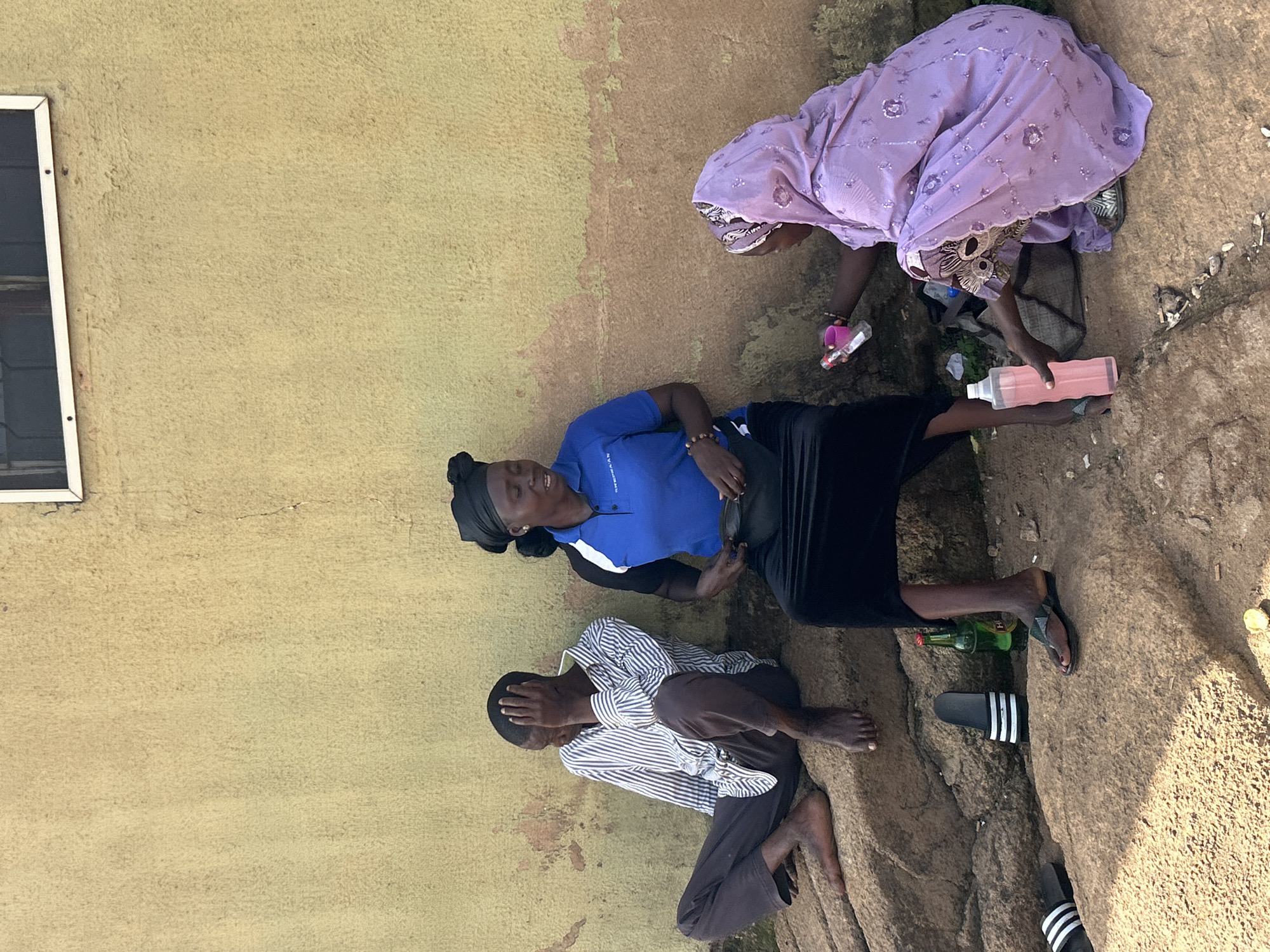 In the city of Jos, Nigeria, three individuals are seated on large, rough rocks against a textured, beige wall. To the left, a person in a pinstripe shirt and dark pants sits with their head in their hand. In the center, a woman wearing a blue polo shirt with a white collar and black headscarf is seated and looking to the right. She has a dark fanny pack around her waist. To the right, a person in a lilac headscarf and patterned robe is kneeling. They are holding a pink bottle and a small pink object. In front of the kneeling person is a large, clear plastic bottle filled with pink liquid. To the right of the seated woman in the blue shirt, a green glass bottle rests on the rocks. Several pairs of flip-flops are visible on the rocks. The ground is rough and rocky with patches of sand. The wall behind the individuals shows signs of weathering and discoloration. A window with a white frame is visible in the upper part of the wall.