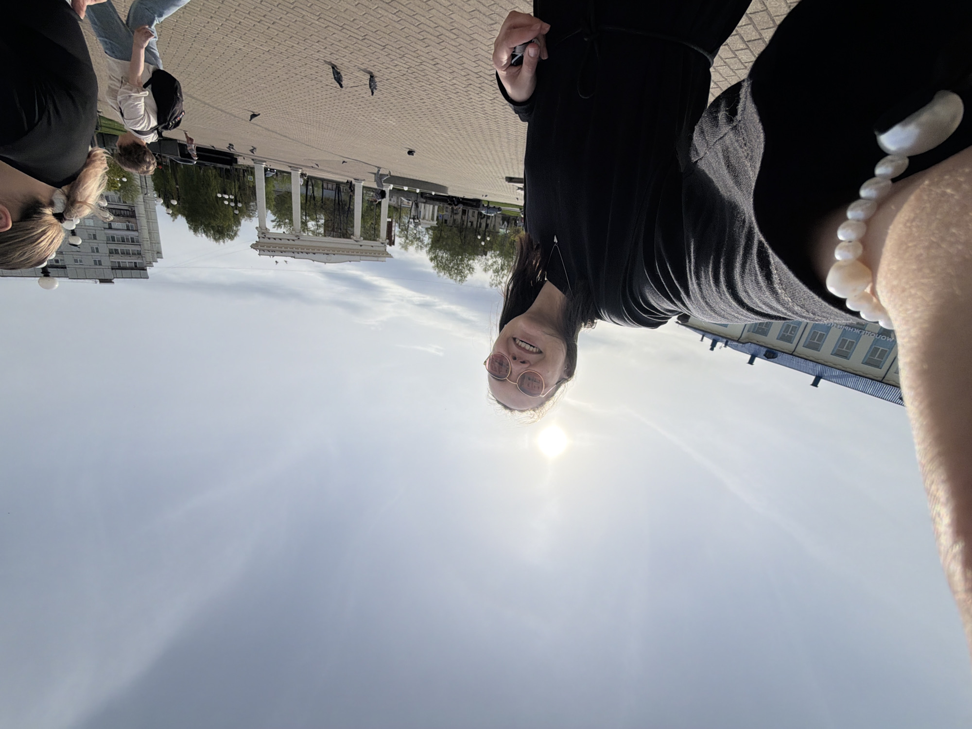 Here is a detailed description of the image:
**Scene:** A selfie taken outdoors in what appears to be the city of Chaikovsky, Russia. The perspective is unusual, with the camera tilted significantly upwards.
**People:**
* A person wearing pink sunglasses is taking the selfie. They are smiling.
* Other people can be seen in the background, walking in the distance.
**Objects:**
* A columned building is visible in the background, likely a recognizable landmark.
* Birds are scattered on the ground.
* A pearl bracelet is on the right arm.
* A dark-colored garment (possibly a dress or a top and pants) is worn by the person taking the photo.
**Actions:**
* The primary action is the person taking a selfie.
* Other people are walking.
**Environment:**
* The scene is set in a public space, possibly a town square or a park.
* The ground is paved with brick.
* The sky is bright.
* There are buildings in the distance.