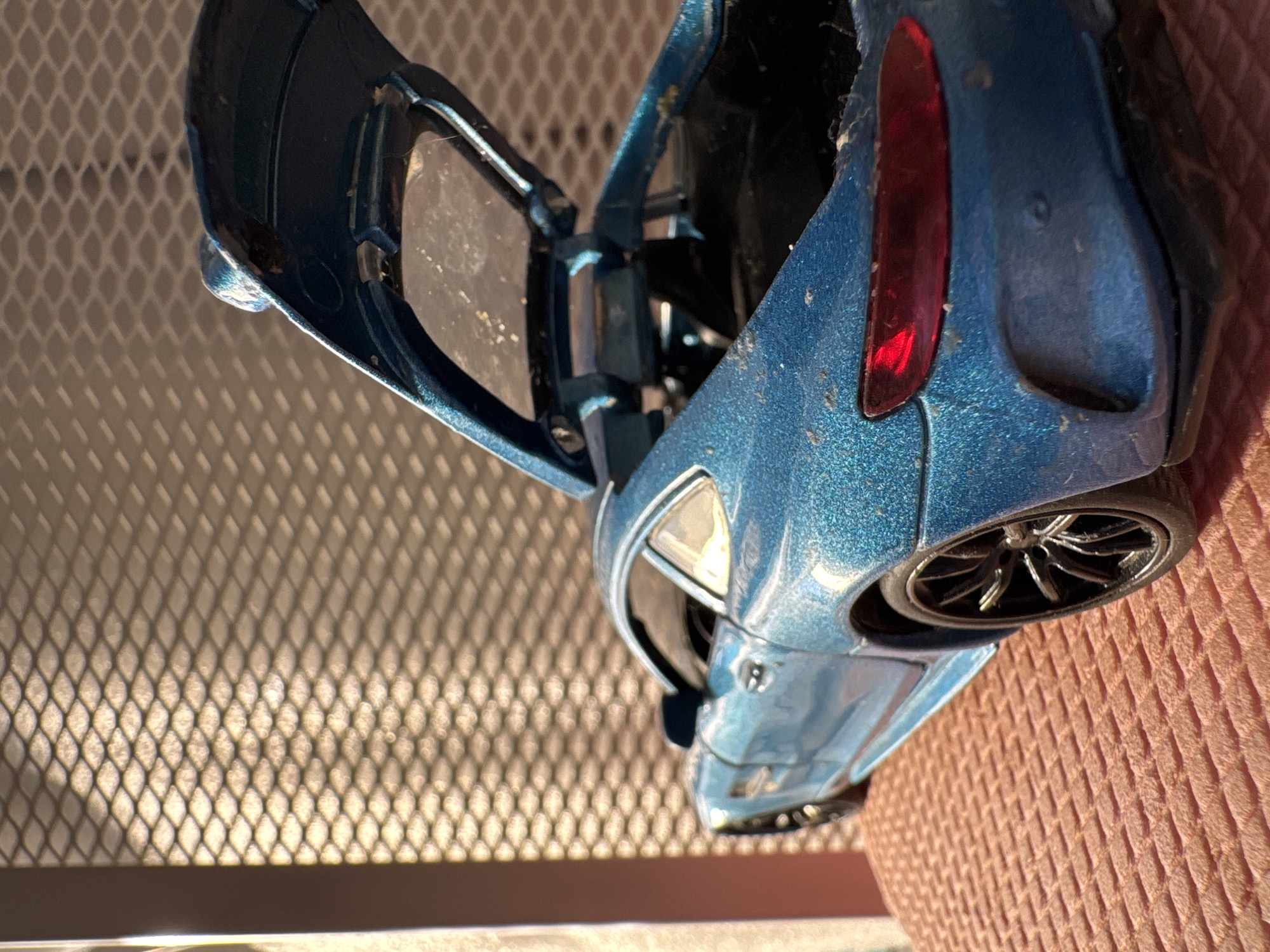 A blue die-cast model car with open gullwing doors rests partially on a pink, textured surface, and partially on a diamond-patterned metal grate. The car is dusty and shows signs of play wear. The location, Novo-Nikol'skoe, Russia, is not discernible from the image alone.