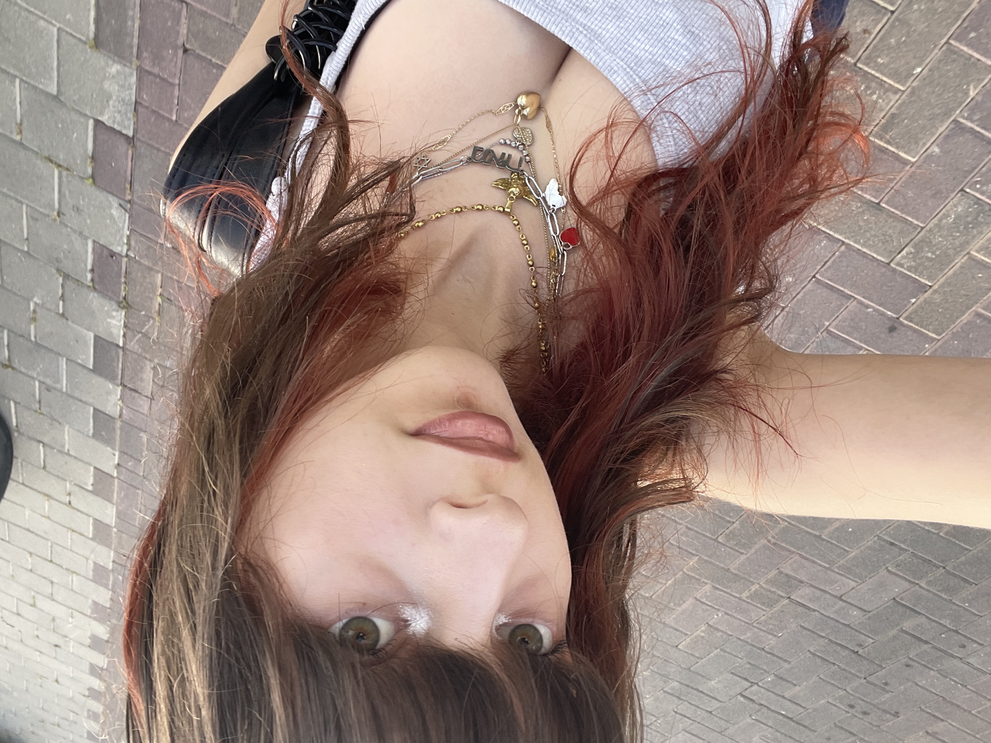 A close-up, overhead selfie captures a young person from an upward-looking perspective, with their head tilted back and face appearing inverted. The subject has medium-length brown hair with prominent red or auburn highlights that frame their face and extend downwards. Their eyes are green or hazel, accentuated by a bright white highlight on the inner corners. Their lips are painted with a muted pink or nude shade.
They are wearing a light-colored, possibly grey, ribbed or knit top. Multiple layered necklaces adorn their neck, featuring various charms. These include a gold chain with a heart pendant, another with a small angel charm, and a silver-toned chain from which a pendant spells out "DALI" or "DAILY". A small red heart charm is also visible on one of the necklaces. A black strap or accessory is partially visible on the left side of the frame.
The background is a grey-toned paved surface with a distinct herringbone pattern, indicating an outdoor setting such as a sidewalk or plaza. The bright lighting suggests it is daytime. The image is a casual self-portrait taken in Raanana, Israel.