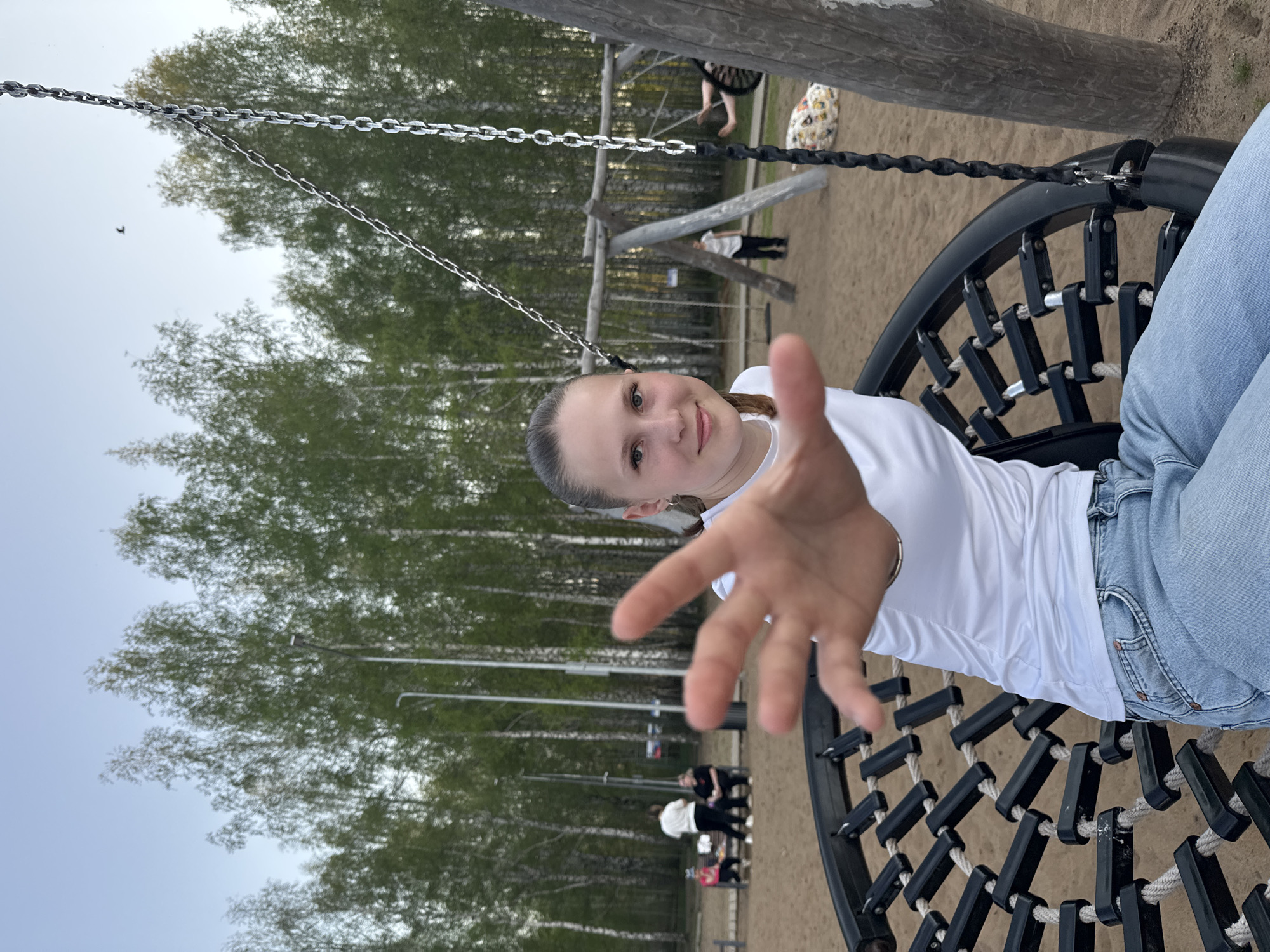 A young woman is centered in a playground. She is smiling and sitting on a swing that is made of a circular black metal frame woven with rope. She extends her right hand towards the viewer. The woman wears a white t-shirt and light blue jeans. In the background are trees and a sandy area. There are other people in the distance. The scene is in the city of Chaikovsky, Russia.