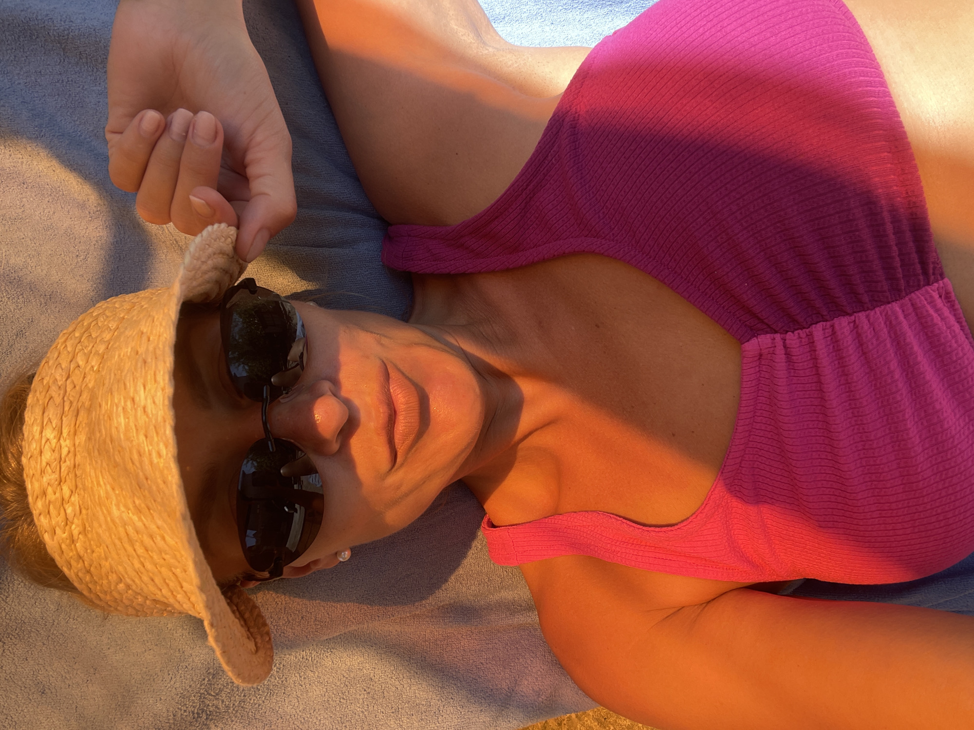 A woman wearing a straw visor and sunglasses is lying down on a blue towel, her body angled towards the right side of the frame. She has on a bright pink, ribbed tank top or bikini top. The sun is casting warm, golden light across her face, shoulders, and arm, creating strong shadows and highlights. Her hand is resting on the brim of her visor. The background consists of the blue towel. The scene appears to be outdoors, possibly at a beach or poolside, during golden hour. The location in Baranovka, Russia is not evident from the image.