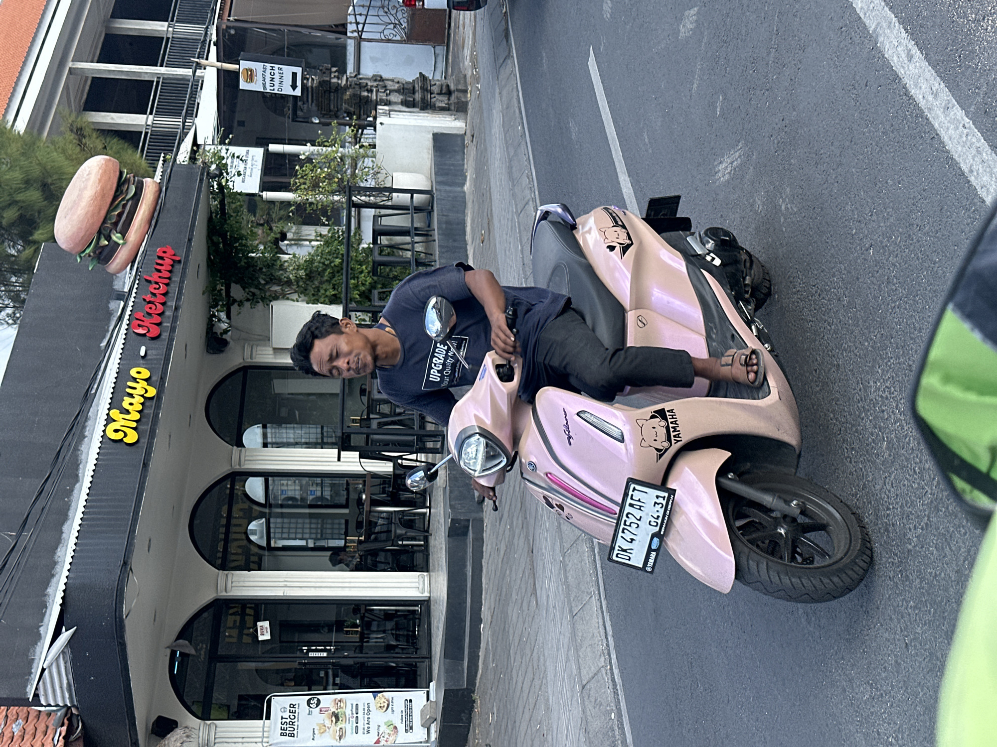A man rides a light pink Yamaha scooter down a paved street in North Kuta, Indonesia. The man is wearing a dark blue t-shirt, black pants, and sandals. He is looking straight ahead. The scooter has a license plate that reads "DK 4752 AFT 04.31." A sticker of a cat wearing headphones is on the front of the scooter. In the background, a building with a sign that reads "Mayo Ketchup" is visible. There is also a large burger statue on top of the building. The street is lined with buildings, including a restaurant with outdoor seating. A sign on the side of the street indicates "Breakfast Lunch Dinner" with an arrow pointing to the right. The weather appears to be clear and sunny.