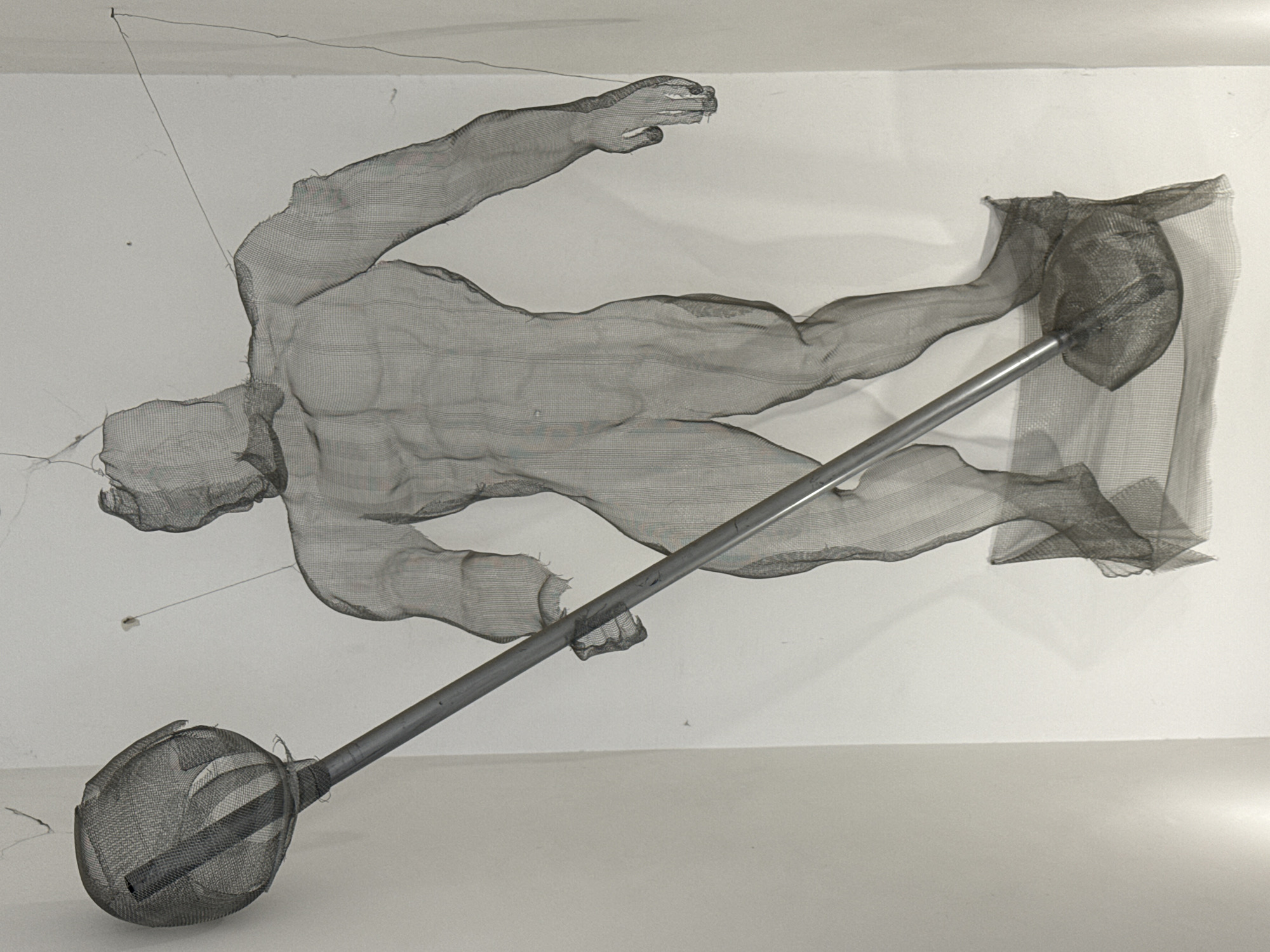 A wire mesh sculpture of a human figure lifting a barbell is suspended in an indoor setting, likely an art gallery or studio, against a white wall and floor. The figure is depicted horizontally, mid-lift. The barbell features wire mesh or netting-encased weights at both ends. Fine wire lines, likely part of the sculpture's suspension system, extend from the figure to the wall and ceiling. The sculpture's location in Varna, Bulgaria is not evident from the provided image.