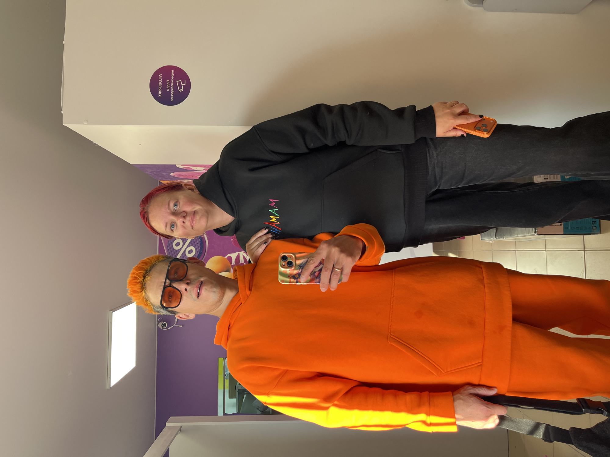 Two individuals are posing for a selfie in a brightly lit, indoor environment. The person on the left has vibrant orange, spiky hair, wears orange sunglasses and an oversized bright orange hoodie and matching sweatpants. They are holding a smartphone in their right hand, which displays an image on its screen and has a patterned phone case. A black strap, likely from a bag, is visible in their left hand. The person on the right has red hair and is wearing a black hoodie with colorful lettering that appears to spell out "HAMAM". They are wearing dark wash jeans and holding an orange smartphone in their right hand. They have their left arm resting on the shoulder of the person on the left.
To the right of the individuals, on a white wall, there is a circular purple sticker with text and an icon. The text on the sticker is in Cyrillic and reads "WILDBERRIES" in an arc and "Всё будет хорошо" below it, which translates to "Everything will be fine." There is also an icon of a security camera. The background includes a purple wall to the left with some blurry graphics, a fluorescent light fixture on the ceiling, and tiled flooring. The overall impression is a casual, perhaps retail or service environment.
The presence of the "WILDBERRIES" branding on the sticker strongly suggests the location is a Wildberries pick-up point or store. Wildberries is a major Russian e-commerce company. Given this information and the context of the branding, the image is likely taken at a Wildberries location in the city of Chaikovsky, Russia.