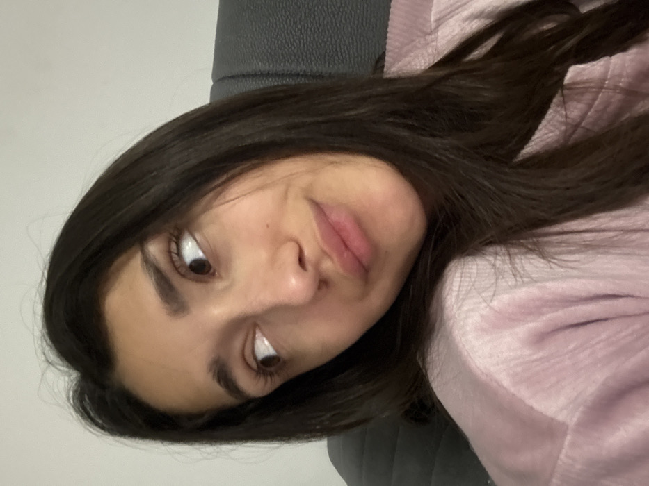 A young woman with long, dark hair tilted her head and looks off to the side with a slightly pouty expression. Her skin tone is light, and she appears to be wearing a light purple garment. A gray headrest is visible behind her head. She has dark eyebrows and eyes and is indoors.