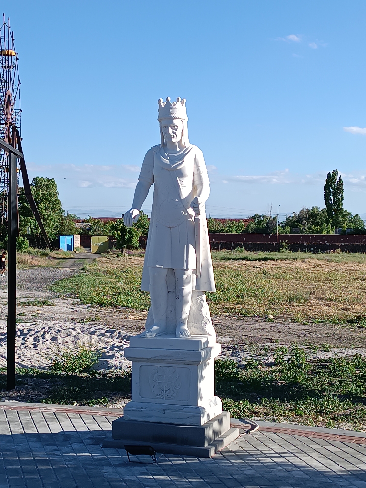 A white stone statue of a king wearing a crown stands on a pedestal in an outdoor setting. The statue is centrally positioned in the frame and faces forward. The king holds an object in his right hand. The pedestal has decorative carvings. In the background, there is dry grass, trees, and a blue sky with some clouds. To the left, a large metal structure, possibly part of a Ferris wheel, is visible. A person wearing dark clothing and a light-colored shirt is walking on a paved path in the far left of the image. The paving stones in the foreground are grey with red and brown accents. The scene appears to be a public area with a monument. The statue and its surroundings do not strongly suggest a specific location in Tallinn, Armenia, as there is no clear visual indicator to confirm this city or country.