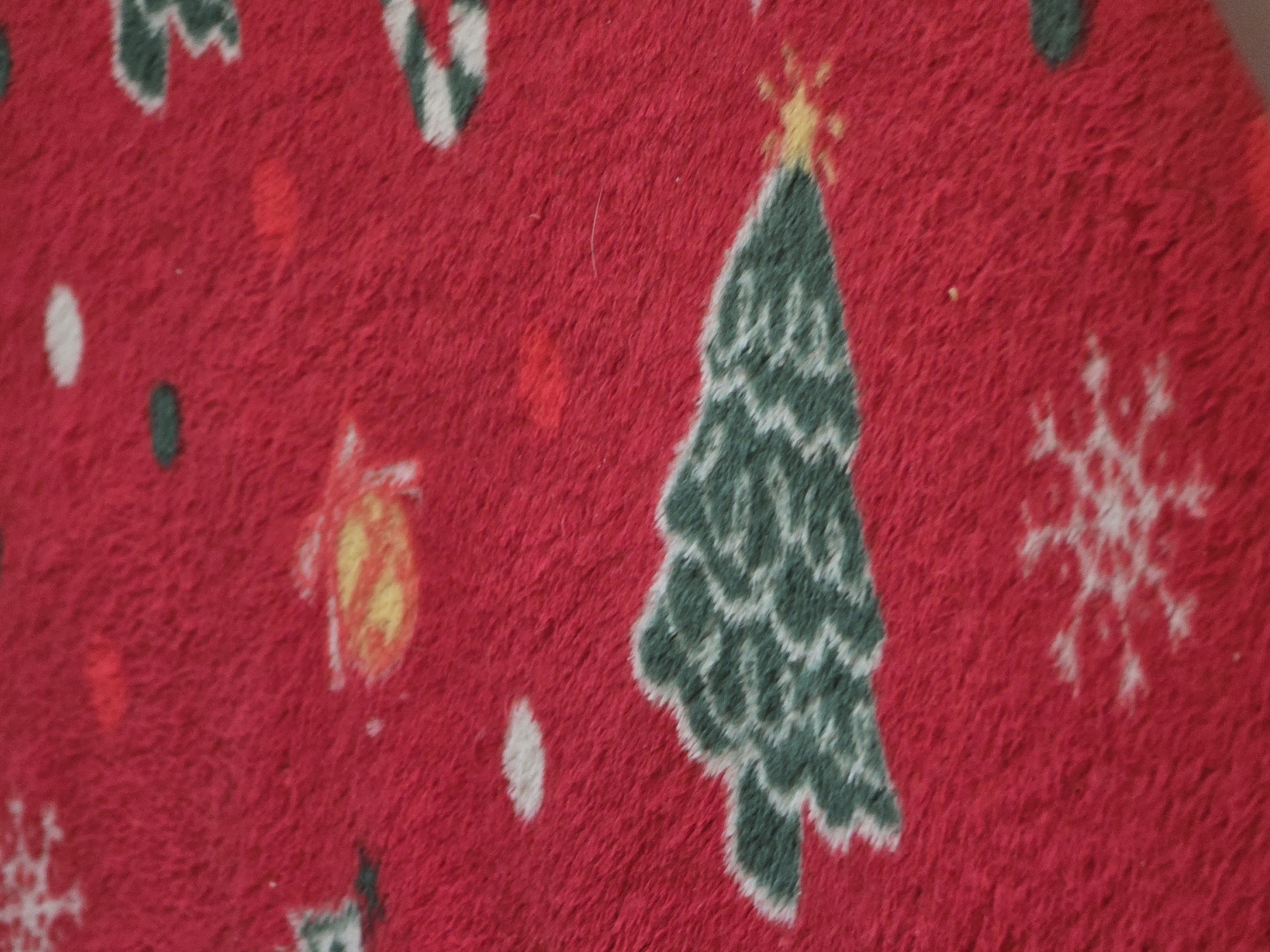 A close-up, overhead shot captures a portion of a red, plush textile, possibly a rug or blanket, adorned with a Christmas-themed pattern. The design features stylized green Christmas trees with white outlines, white snowflakes, and scattered white and yellow circular motifs representing snow or ornaments. The texture of the fabric is evident, with visible fibers creating a soft, fuzzy appearance. The image is devoid of any human presence or activity, and no specific geographical markers are present to indicate a location in Golyanovtsi, Bulgaria, or any other city. The scene is static and focuses solely on the decorative textile.