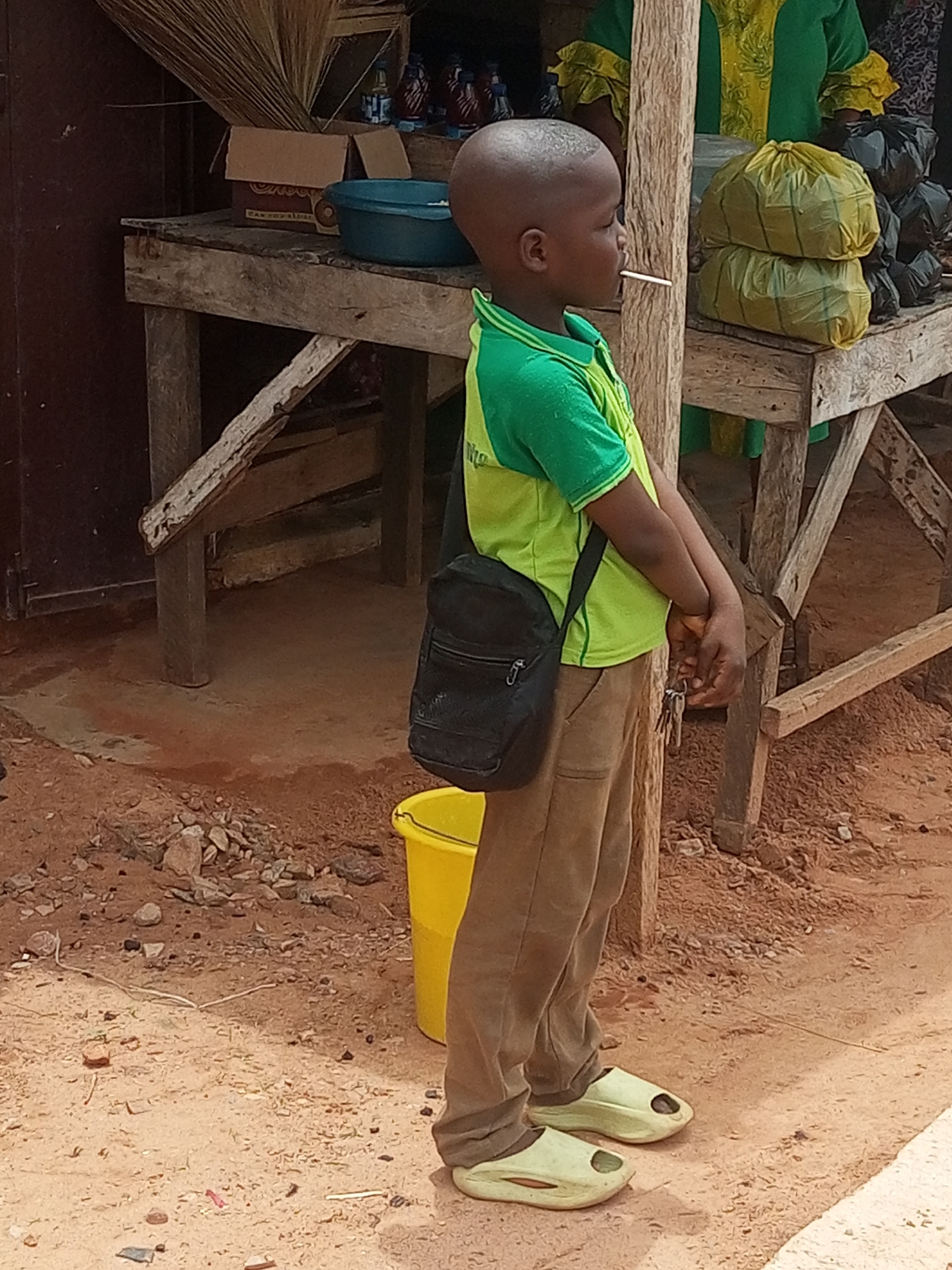 The image depicts a young, dark-skinned boy standing on a reddish-brown, unpaved surface in the city of Unknown, Unknown. He wears a green and yellow short-sleeved polo shirt, light brown pants, and light green slip-on shoes, with a black shoulder bag draped across his body. A white stick-like object is held in his mouth, and he holds a set of keys with clasped hands in front of him, gazing towards the right side of the frame. Behind him, a rustic wooden stall or table is laden with various items: a brown cardboard box, a blue plastic bowl, two stacked yellow plastic bags, and several black plastic bags. Dark-colored bottles are visible on the stall surface to both the left and right. Above the stall, a dense cluster of light brown, straw-like material forms an overhead structure. A partially visible adult, clothed in a green and yellow patterned garment, stands behind the stall. The ground in the foreground consists of dirt, scattered small rocks, and debris, with a yellow plastic bucket positioned near the boy's left foot.