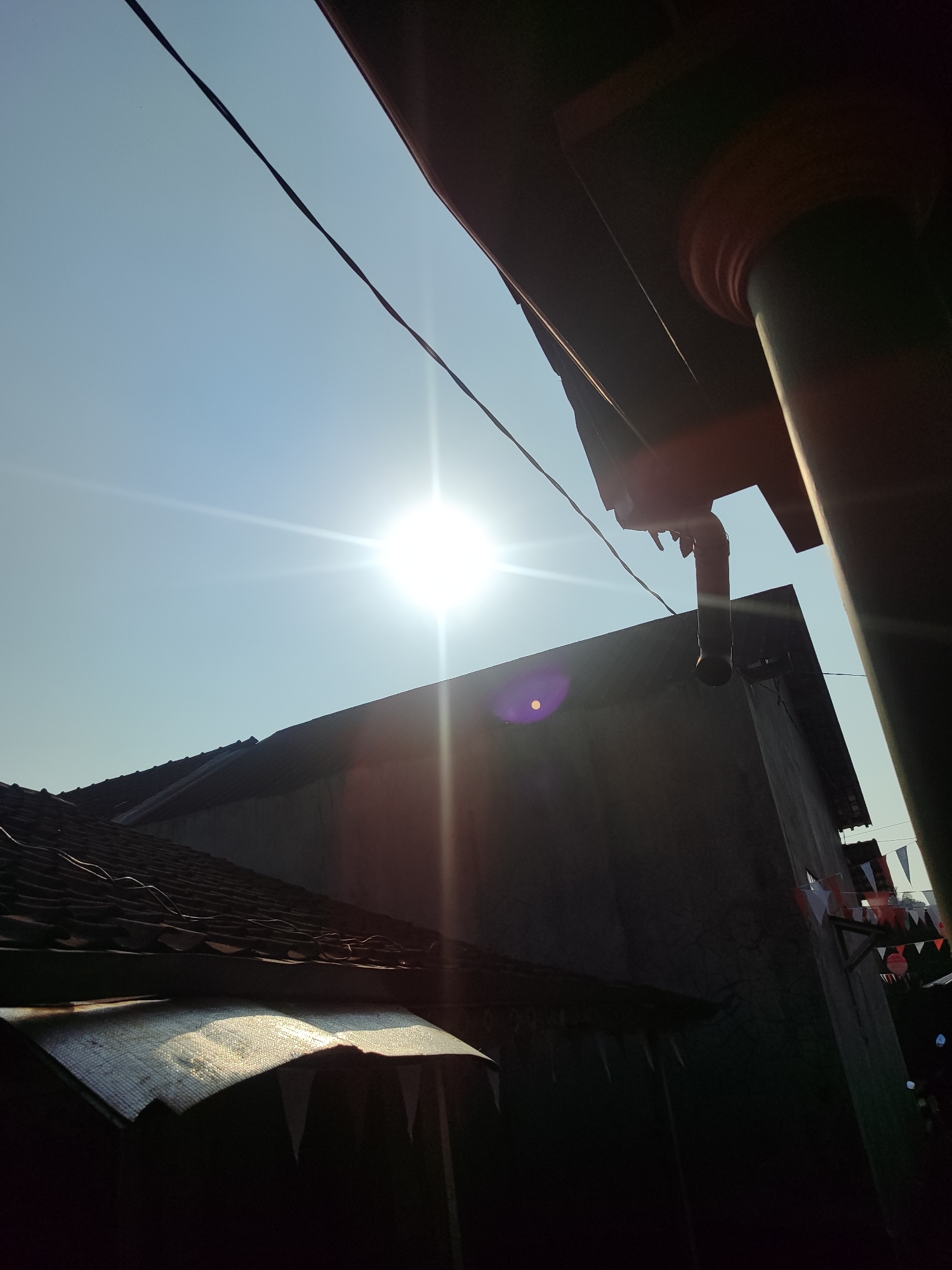 The image depicts an outdoor scene with a bright sun in a clear blue sky. In the foreground, there is a roof made of clay tiles. To the right, a portion of a building structure with a dark pillar and overhang is visible. Electrical wires cross the sky. The overall impression is of a sunny day in a residential or semi-urban area. There are no people or animals visible in the image. The location is identified as Gantungan, Indonesia.