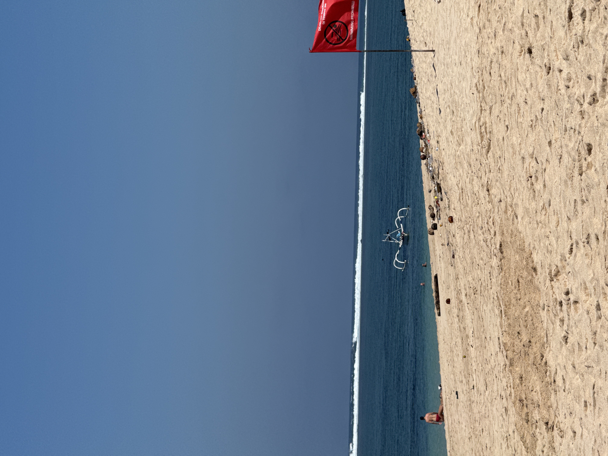 The image shows a wide, sandy beach under a clear blue sky. In the foreground, the sand is textured with footprints and some scattered debris, including logs and trash. Further back, where the sand meets the water, there are more rocks and trash visible. The ocean is a deep blue with white waves breaking near the shore. A traditional Balinese fishing boat with outriggers is visible in the water.
On the beach, a person wearing red shorts is sitting in the sand, facing away from the camera, towards the ocean. To the right of the frame, a red flag is planted on a pole. The flag displays a white circle with a diagonal line through it, and text that reads "DILARANG BERENANG DI TEMPAT INI" and "SWIMMING PROHIBITED". The scene appears to be a public beach in Kuta Selatan, Indonesia, likely one where swimming is not advised due to currents or other hazards. The overall impression is a beautiful natural environment marred by pollution.