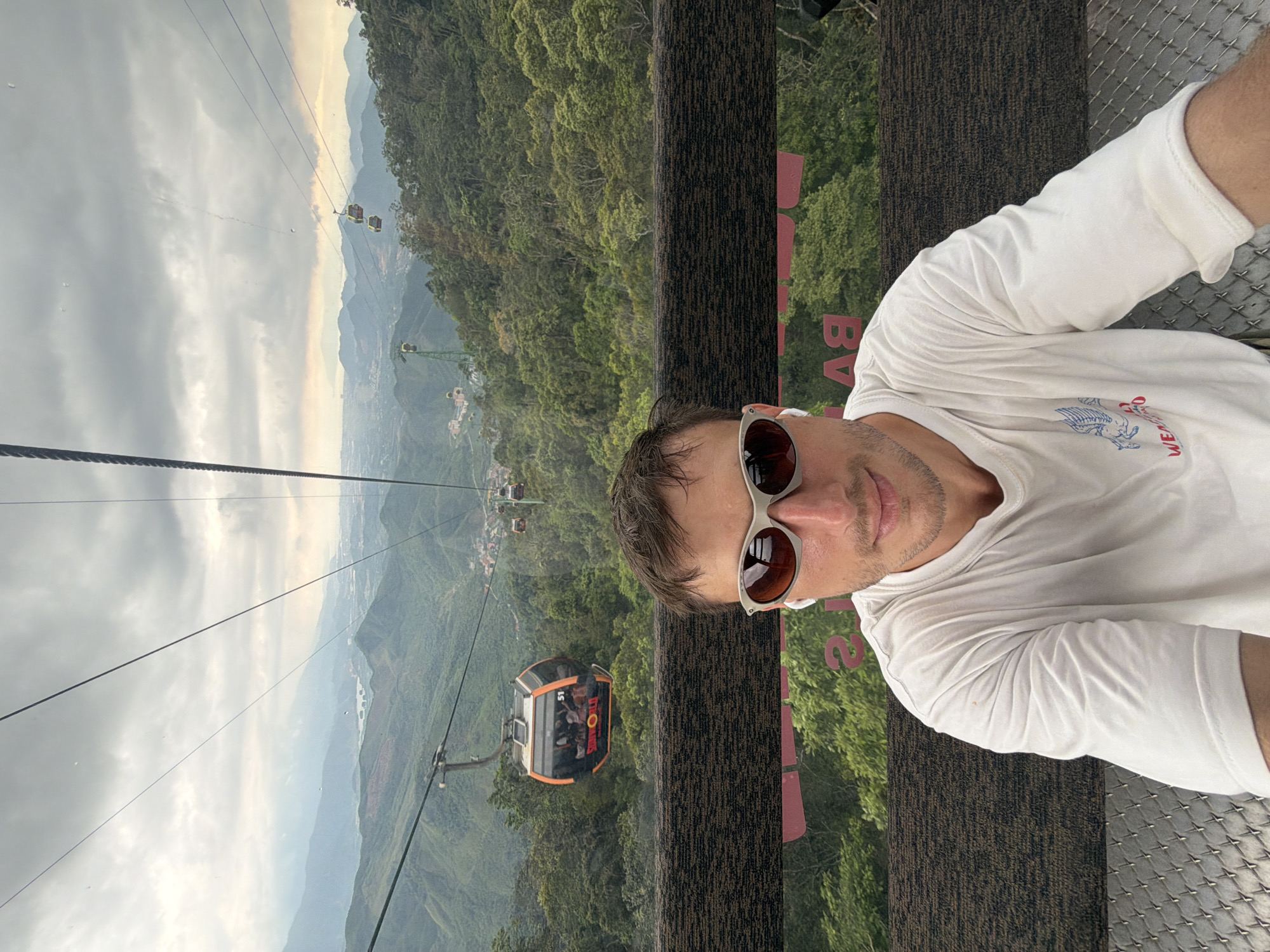 A man is taking a selfie inside a Ba Na Hills cable car near Đà Nẵng, Vietnam. He is wearing sunglasses and a white long-sleeved shirt and has earbuds in his ears. Outside the cable car, a valley filled with green trees and other cable cars on lines is visible. The sky is cloudy. The interior of the cable car has a patterned wall and metal mesh flooring.
