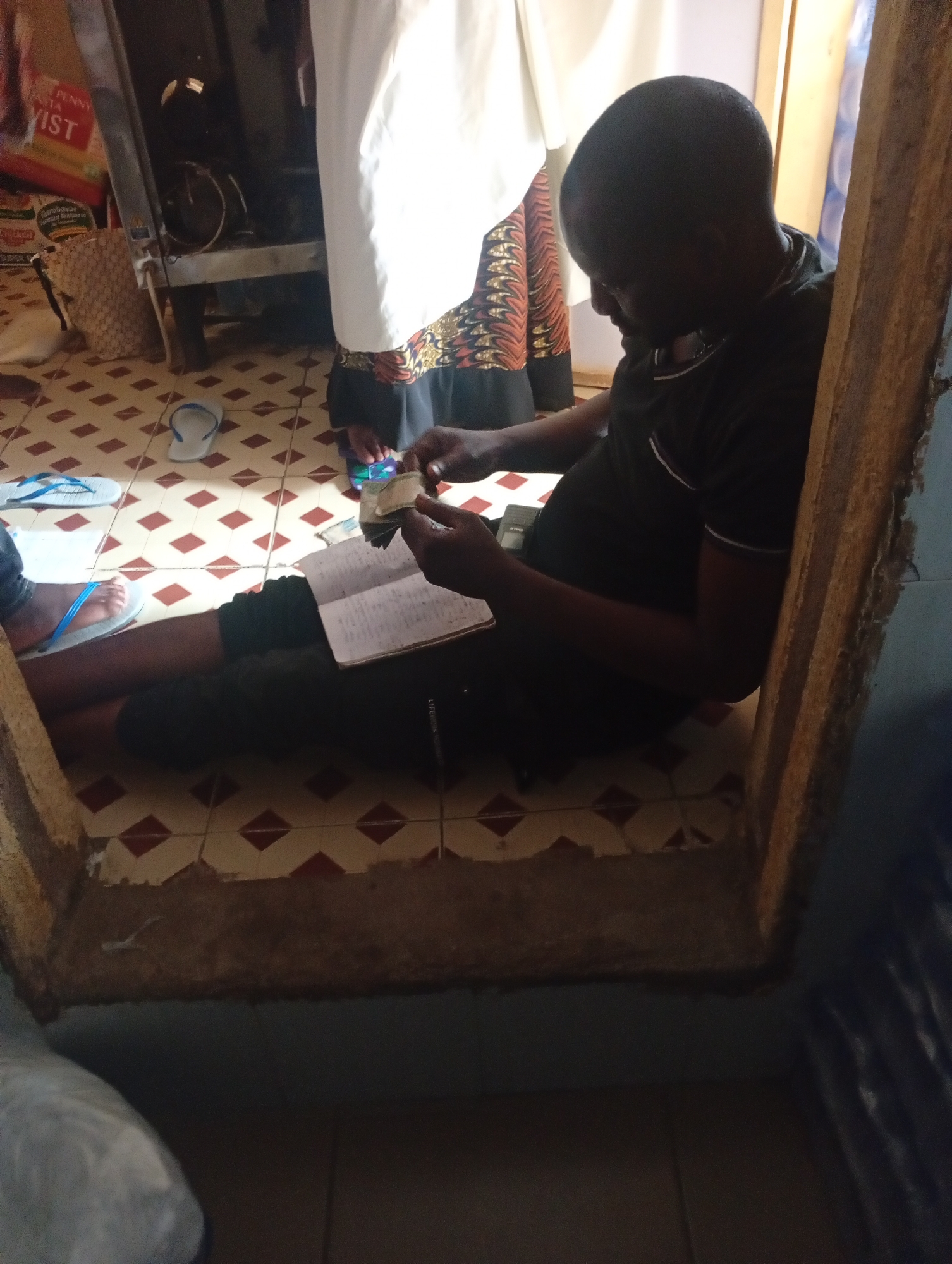A dark-skinned male in a black t-shirt is sitting on a tiled floor, looking down at a notebook and a stack of cash he is holding in his hands. He is leaning against a wooden frame. The floor has a diamond pattern of red and beige tiles. In the background, to the left, is a white curtain and a large appliance. A basket is visible near the appliance. To the right of the frame, a blue tarp or plastic sheeting is visible. The scene appears to be indoors in a simple dwelling. The provided context suggests the location is Rafin Par, Nigeria.