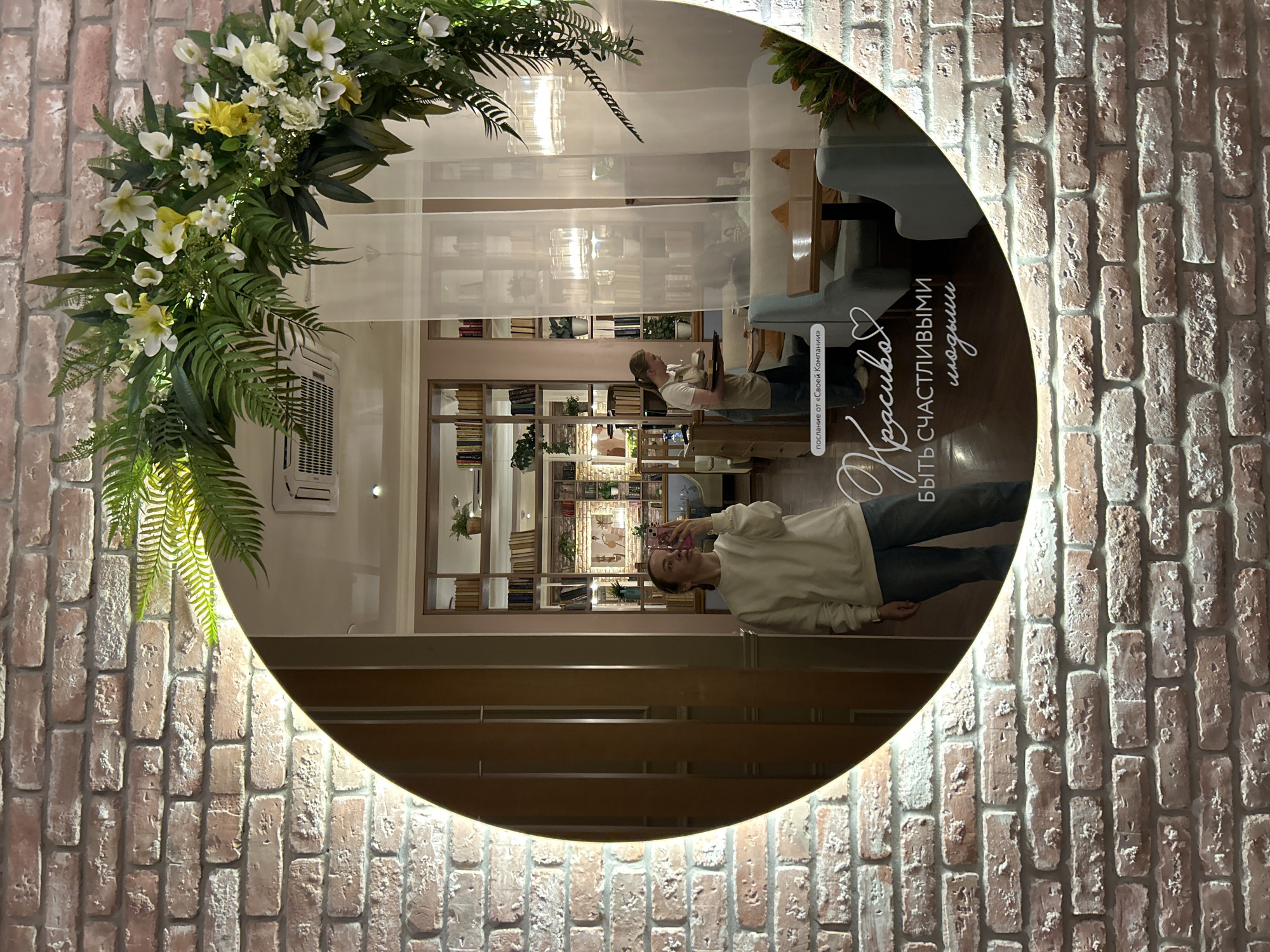A full-shot, eye-level indoor medium, taken in a cafe. A young woman is standing in the foreground, reflected in a large, circular mirror. She is holding a pink smartphone and taking a selfie. She has blonde hair pulled back and is wearing a white sweatshirt and blue jeans. The mirror is decorated with a large floral arrangement of green ferns and white and yellow flowers. The background of the mirror shows a cafe interior with tables, chairs, and bookshelves. The text "Послание от «Своей Компании»" and "Красиво, быть счастливыми людьми" is superimposed on the mirror. In the background, a cafe worker in an apron carries a tray of drinks. The cafe walls are made of brick. The city of Yekaterinburg, Russia, is implied by the phrase "Своей Компании" which is a reference to a chain of restaurants popular in that region.