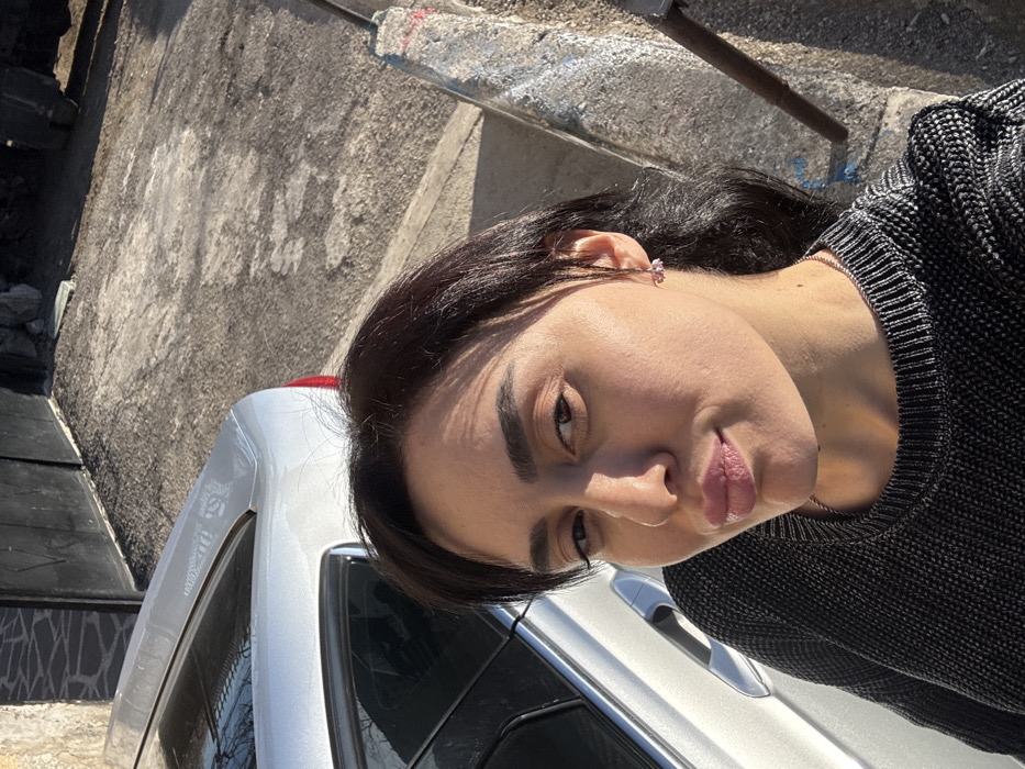 An outdoor, eye-level photograph captured in Bolnisi, Georgia, depicts a single female subject positioned centrally, facing the camera. She has dark hair, dark eyebrows, and wears a dark grey knitted sweater and light-colored earrings. Her expression is a slight pout with direct eye contact. Sunlight illuminates the right side of her face and body, casting shadows on the left.

Behind her, to the left, is the rear quarter panel and trunk of a light-colored (possibly silver) sedan, with a portion of the rear window and red taillight visible. The ground immediately behind the car is an uneven, dark-colored paved surface, possibly asphalt or concrete. Further left and behind the vehicle, a light-colored wall with visible texture is present, adjacent to a dark, possibly metal gate or door.

To the right of the subject, a weathered concrete curb or low barrier, marked with faint red and blue paint, runs vertically. A dark, thin pole or rod ascends from behind this curb. The overall scene suggests a residential or urban outdoor setting under direct sunlight.