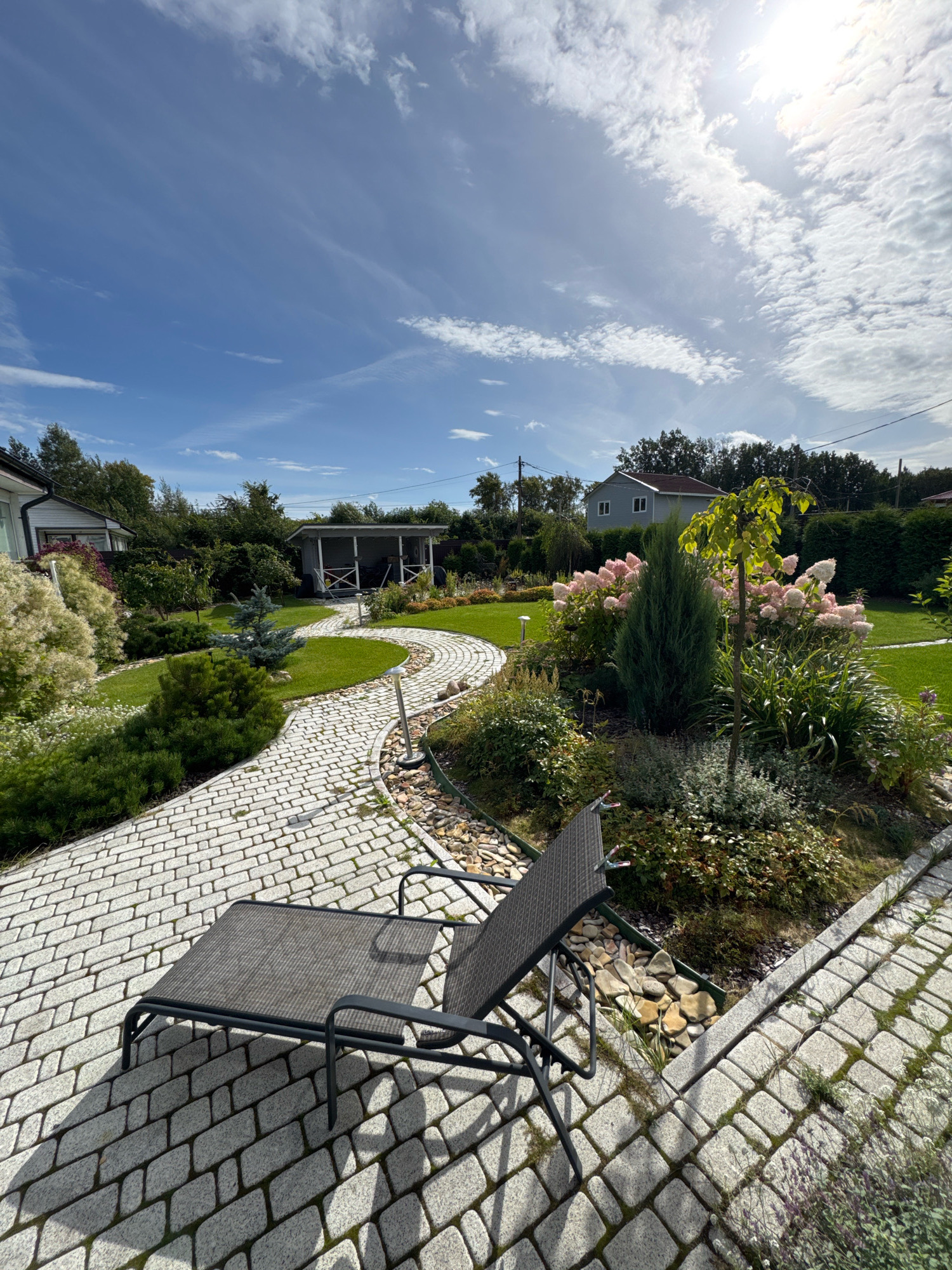 This image depicts a well-maintained garden in Oliki, Russia. A grey chaise lounge is positioned on a winding pathway made of light-colored cobblestones. The pathway leads through a garden filled with various plants, including flowering shrubs with pink blooms, evergreen trees, and other ground cover. In the background, a small white gazebo with an open structure is visible, and behind that, a light blue two-story house with a dark roof. The garden is bordered by a dense green hedge and mature trees. The sky above is blue with scattered white clouds, and the sun is shining brightly. The overall scene conveys a sense of tranquility and order.