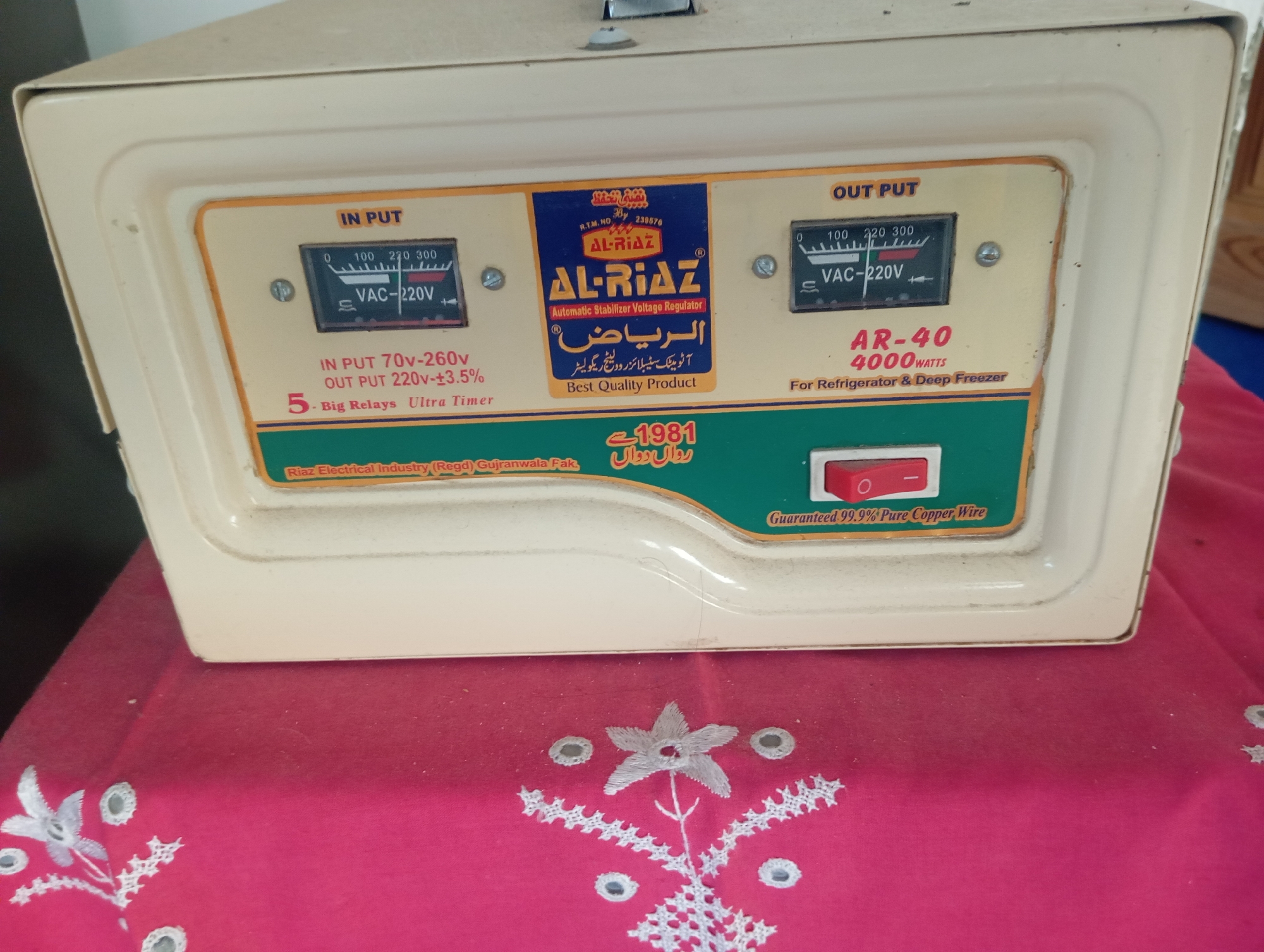 The image shows a close-up of a cream-colored voltage stabilizer, likely manufactured by "Al-Riaz." The device is placed on a pink fabric tablecloth adorned with white floral embroidery and small mirrors. The stabilizer features two analog voltmeters, one labeled "IN PUT" and the other "OUT PUT," both indicating voltage readings. Text on the stabilizer includes "Automatic Stabilizer Voltage Regulator," "AR-40 4000 WATTS," and "For Refrigerator & Deep Freezer." There's also a red on/off switch. The lower section of the stabilizer displays text in Urdu, which translates to "Riaz Electrical Industry (Regd) Gujranwala Pak." and the year "1981." The presence of "Gujranwala" in the text suggests a connection to that city, not necessarily Dangari.