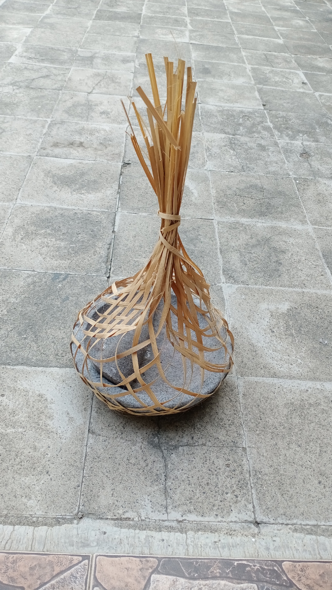 An overhead medium shot captures a natural woven basket filled with stones, resting on a tiled floor. The basket, made from dried bamboo strips, is secured at the top with a tighter weave. The stones are grey and varied in size, partially visible through the open weave of the basket. The tiled floor is composed of square grey tiles with visible grout lines, suggesting an outdoor setting. The lighting indicates a bright day, casting soft shadows. There are no people or animals present in the image. The scene appears to be in a residential area in Mertan, Indonesia, as evidenced by the typical outdoor tiling and natural materials used in the basket's construction.
