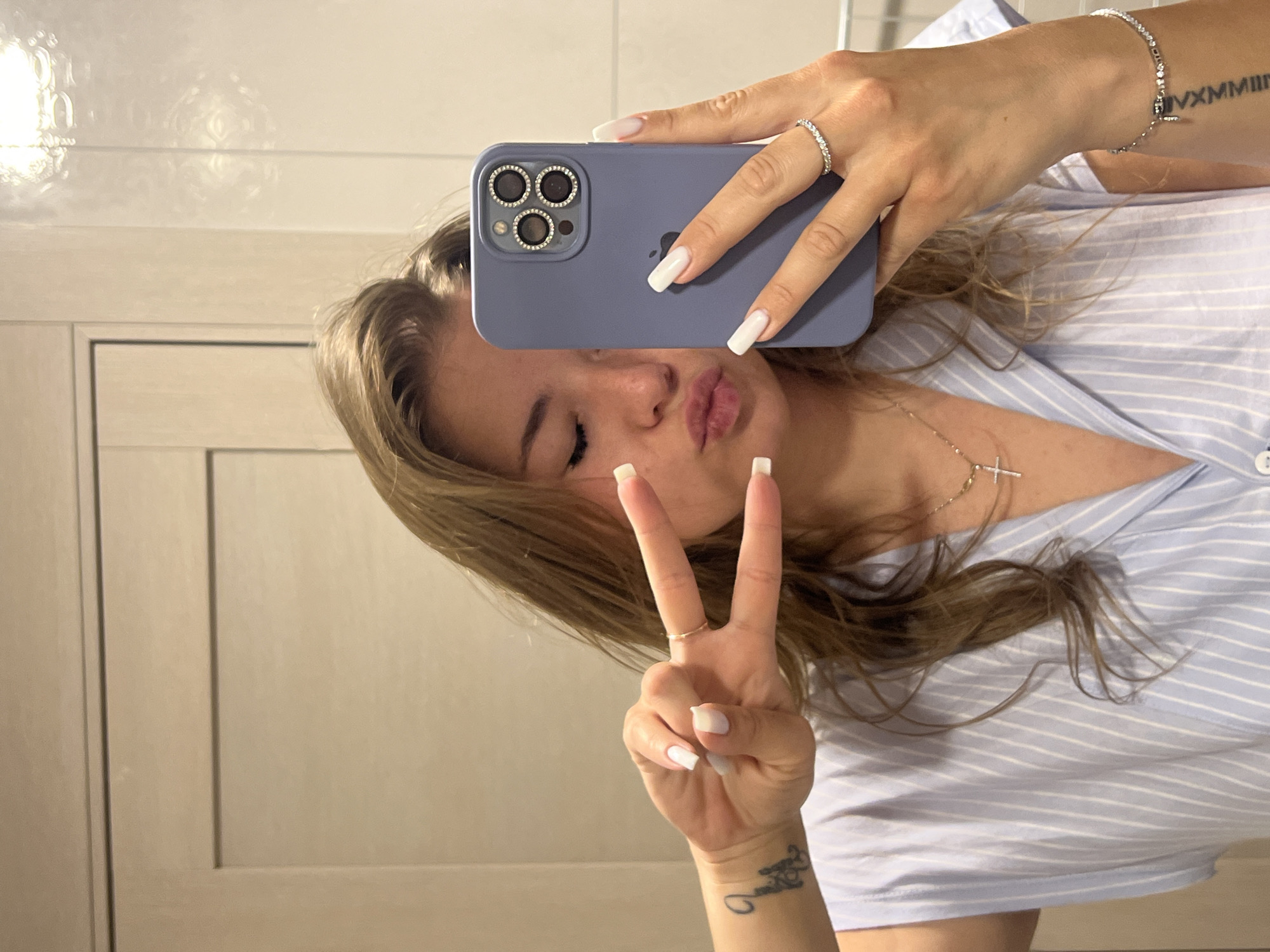 A young adult female is depicted in a mirror selfie, captured from the waist up in a close-up perspective. She has light brown, wavy hair. Her eyes are closed, and her lips are pursed. She is wearing a light blue and white striped sleeveless button-up top. Her left hand is raised, forming a peace sign, displaying a thin gold-colored ring on her ring finger and a small, dark tattoo resembling a stylized butterfly on her outer forearm. Her right hand holds an iPhone, equipped with a light purple silicone case and decorative, possibly jeweled, rings around its three camera lenses, obscuring the right side of her face. A silver-colored ring with small stones is visible on her right ring finger. A thin gold-colored chain with a small silver-colored cross pendant hangs around her neck. On her left wrist, a silver-colored bracelet with multiple small stones is worn, and Roman numeral tattooing is visible on her inner forearm. The background consists of light beige-colored walls, appearing to be tiled on the right side, and a light-colored door or cabinet frame on the left. A blurred reflection of an overhead light fixture is visible in the upper right portion of the background. The scene is an indoor domestic or hotel setting, consistent with a bathroom or dressing area. No specific visual cues within the image identify a precise location in the city of Budva, Montenegro.