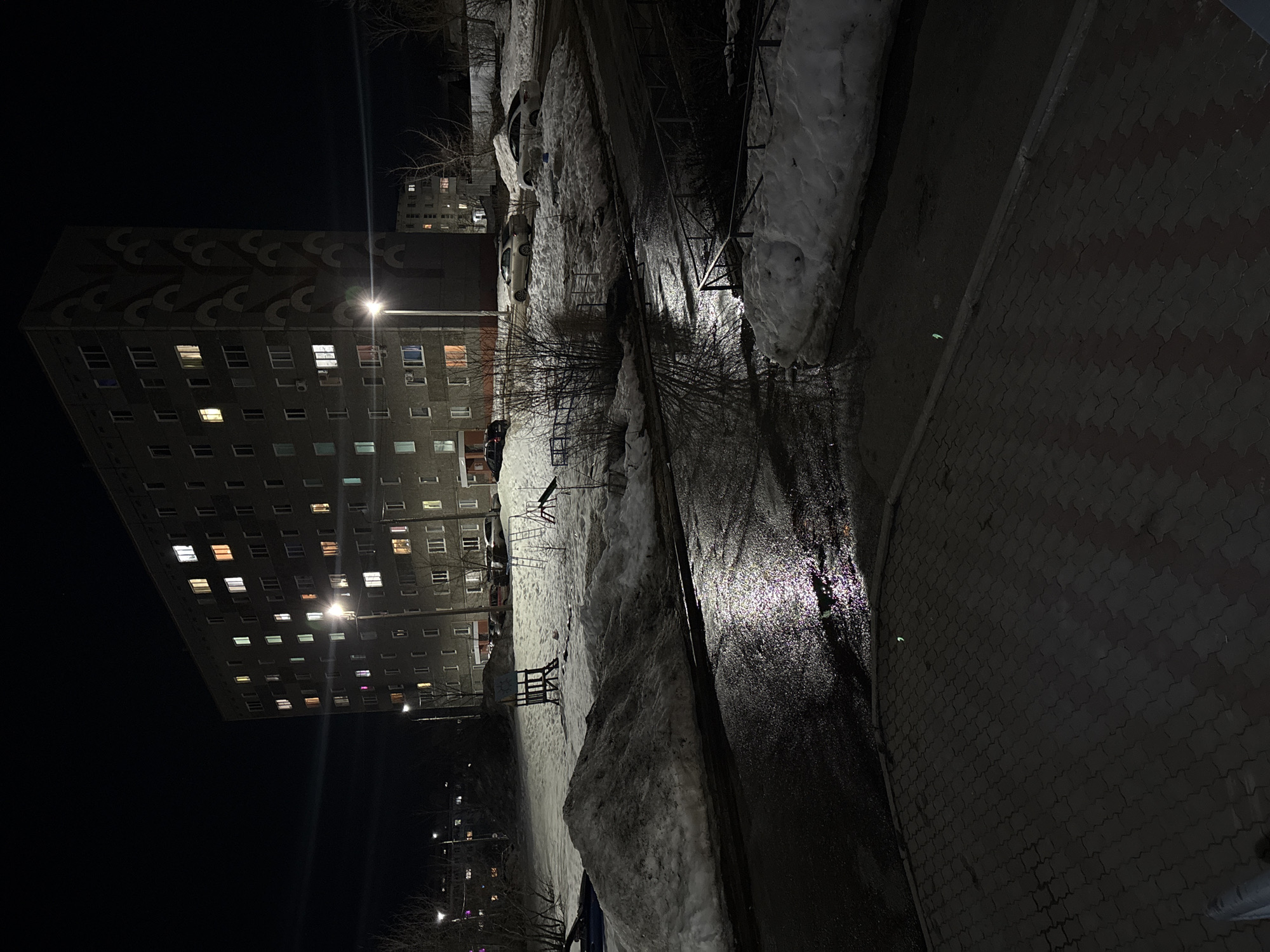 The image depicts a nighttime scene in a residential area of Chaikovsky, Russia, characterized by artificial illumination and significant snow cover.
In the center and left background stands a dark grey, multi-story (approximately 9-10 floors) concrete apartment building, likely a dormitory or block of flats, with numerous windows. Many windows are lit, emitting various hues of light. The building's right-side facade features a large, stylized, relief-like pattern resembling a stacked, repeating wave or tree motif in a darker grey or brown. Two bright streetlights illuminate the immediate vicinity of this building; one is centrally positioned, causing a lens flare effect, and the other is further left.
The ground comprises a wet asphalt road or pathway in the midground, reflecting the streetlights. This wet surface separates a dark, patterned pedestrian paver area in the foreground from snow-covered terrain. Large, irregular piles of snow, some appearing dirty, dominate the midground and right side, partially obscuring a snow-covered playground area containing a dark wooden structure (possibly a tower or slide base) and a lighter metal climbing frame. Several parked cars, primarily light-colored sedans, are visible behind the snow piles and in front of the main building. Bare deciduous trees and sparse bushes are scattered throughout the scene, consistent with a winter or early spring environment. A darker, less distinct residential building with illuminated windows is visible in the far background on the right. A dark metal railing is partially visible on the right edge of the foreground, bordering a snow drift. No individuals are present.