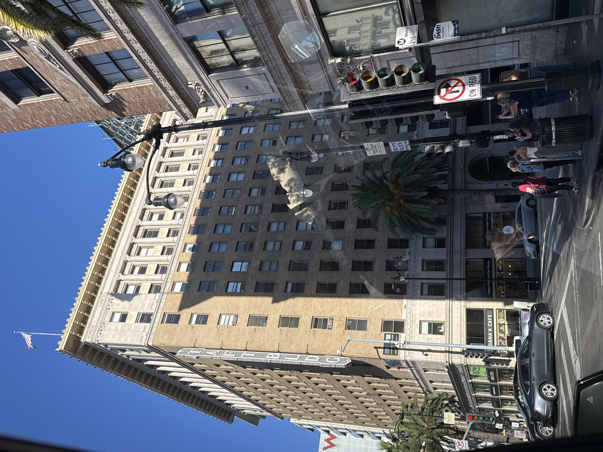 The image captures a street view in Los Angeles, California, USA. A prominent brick building, identified by a vertical sign as the "TAFT BLDG," dominates the center of the frame. Its facade features numerous windows and ornate architectural details along the roofline. In front of the Taft Building, a palm tree adds a tropical element to the urban setting.
On the right side of the image, another building, constructed of brick and stone, is partially visible. A traffic light system is positioned on the sidewalk, with a "No Turn on Red" sign prominently displayed. Several people are standing on the sidewalk, some holding cell phones as if taking pictures. Cars are seen on the street, with one gray sedan in the foreground. The sky is a clear, bright blue, indicating a sunny day.
In the background, a taller, modern building with a distinctive red "W" logo is visible. Other street signs, including one for "Metro 210" and "DASH HOLLYWOOD," indicate the location is a public transportation hub. The overall scene depicts a bustling urban environment with a mix of historical and modern architecture.