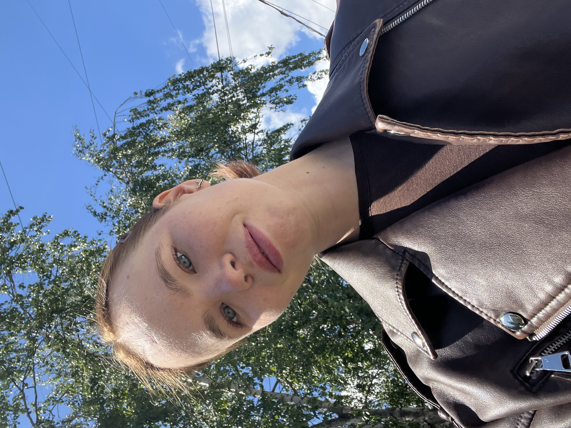A woman with light skin and blue eyes wearing a brown leather jacket takes a selfie from a low angle. A tree with green leaves and a blue sky with clouds are in the background. Overhead power lines are also visible. The location is possibly in the city of Chaikovsky, Russia.