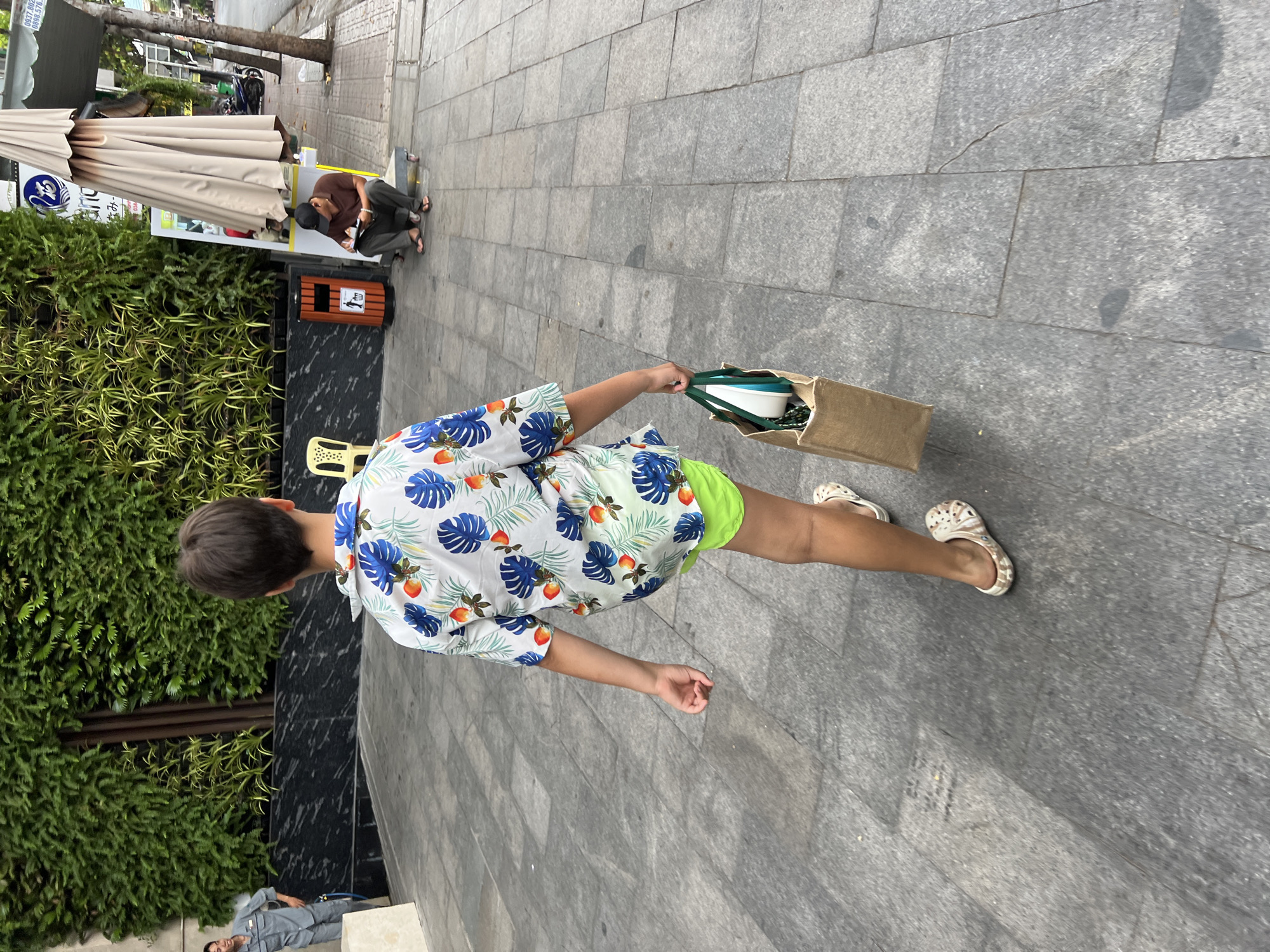 A young person, seen from behind, walks on a tiled sidewalk in Nha Trang, Vietnam. They are wearing a white short-sleeved shirt with a blue leaf and orange fruit pattern, lime green shorts, and patterned clogs. The person is carrying a tan canvas tote bag with green handles in their right hand, which contains a white object and green straps. To the left, a man in a gray uniform stands with his hands on his hips. Further down the sidewalk, a person wearing a dark brown t-shirt, dark pants, and a black baseball cap sits on the ground, writing in a notebook. A large beige umbrella stands behind them, and a brown trash can with a white bin symbol is next to the sitting person. The background features a wall covered in lush green foliage and a street with parked motorcycles and trees.