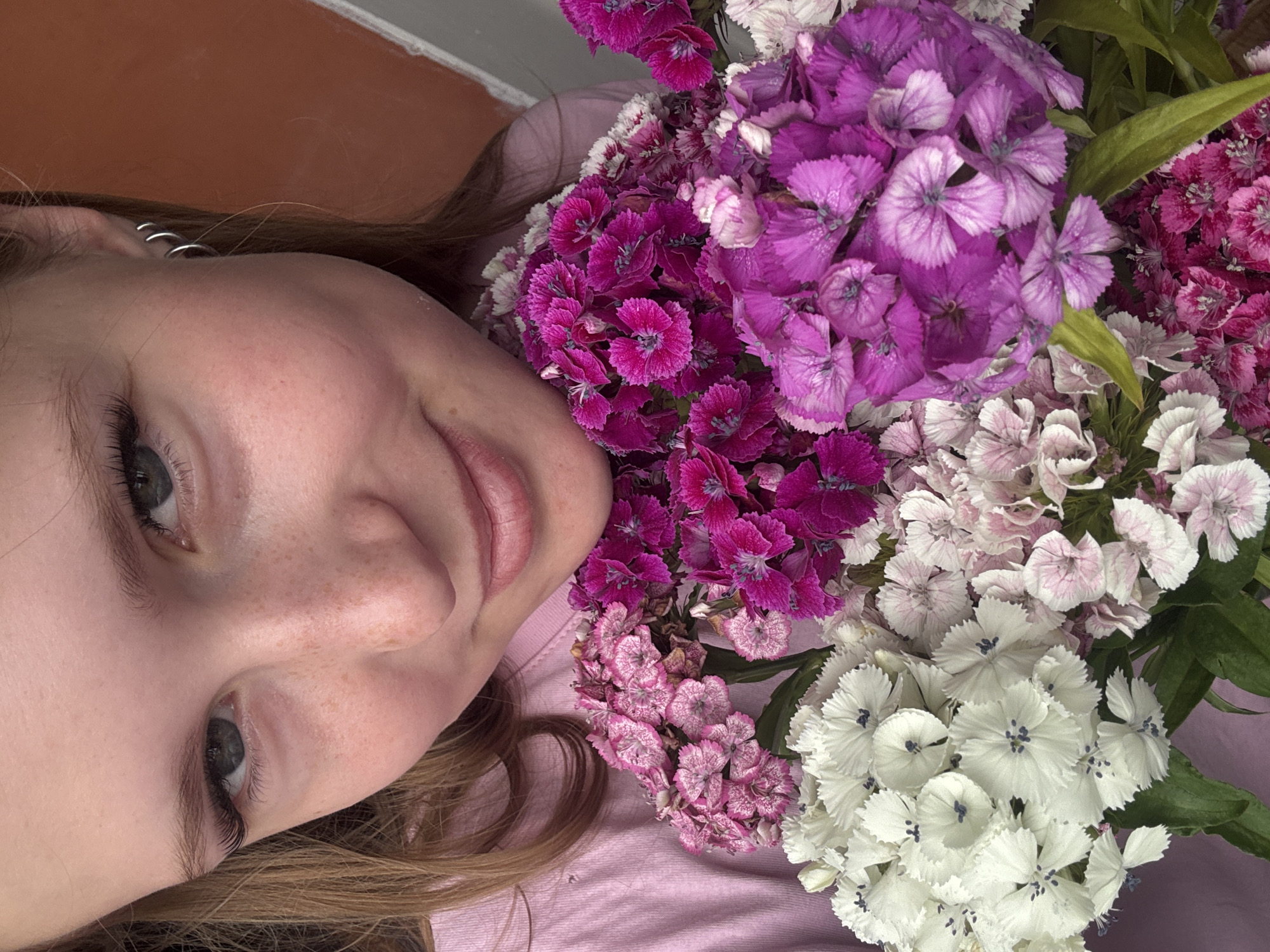 A young woman with blonde hair, freckles, and subtle makeup is holding a bouquet of colorful flowers. She is wearing a pink shirt and has a silver hoop earring in her right ear. The flowers are a mix of white, pink, and purple sweet william (Dianthus barbatus). The background is out of focus, but appears to be a textured orange wall. The image is cropped tightly, focusing on the woman's face and the flowers. The scene does not provide any specific information about its location within the city of Chaikovsky, Russia.