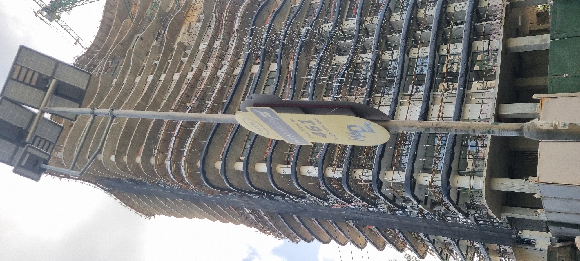The image shows a partially constructed high-rise building in Falomo, Nigeria. The building's architecture is characterized by curvilinear floor plates with protruding structural elements and scaffolding. The façade is currently open, with visible construction materials. A construction crane is visible in the background. In the foreground, a sign, possibly related to a telecommunications provider (MTN), is affixed to a pole. The sky is overcast. There is no visible human activity in the immediate frame.