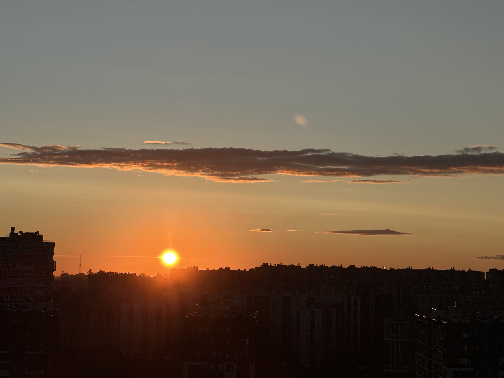 A wide, panoramic, low-angle shot captures a city skyline at sunset. The sun, a brilliant white orb, is partially obscured by a dark silhouette of buildings, casting a warm orange glow across the sky. The sky itself transitions from a pale yellow at the horizon to a soft blue higher up, with streaks of pink and orange clouds dotting the horizon. Several buildings, varying in height and architectural style, are visible in the foreground and middle ground, their dark forms contrasting with the bright sky. The overall impression is one of urban tranquility as day gives way to night.