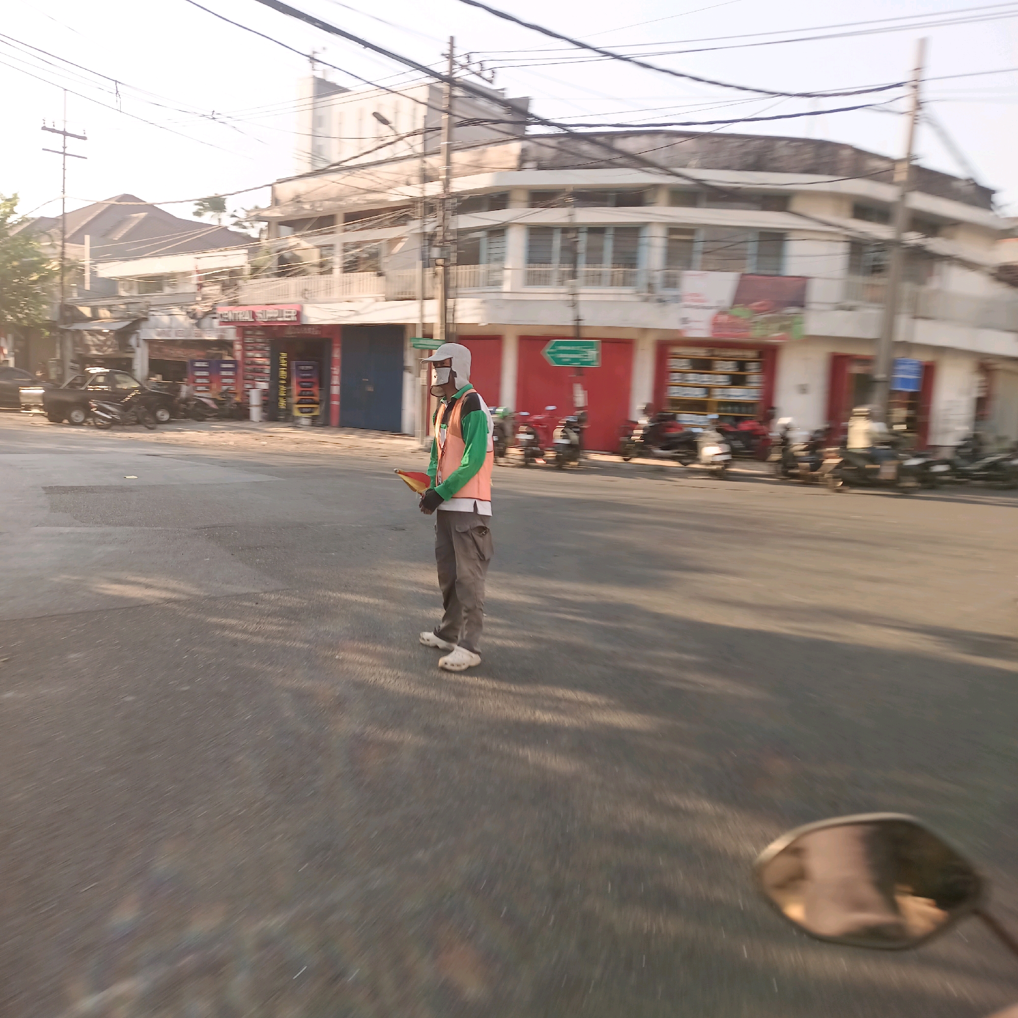 A person wearing a high-visibility orange vest over a green long-sleeved shirt, grey pants, a white helmet, and white shoes stands in the middle of a street in Surabaya, Indonesia. The person is holding a yellow and red flag. The background features buildings with red and white facades, motorcycles parked in front of the buildings, and power lines crisscrossing overhead. The lighting suggests it is daytime, possibly early morning or late afternoon, with sunlight casting shadows on the road. The image appears to be taken from a moving vehicle, indicated by the motion blur and the reflection in the lower right corner, which resembles a motorcycle's side mirror.