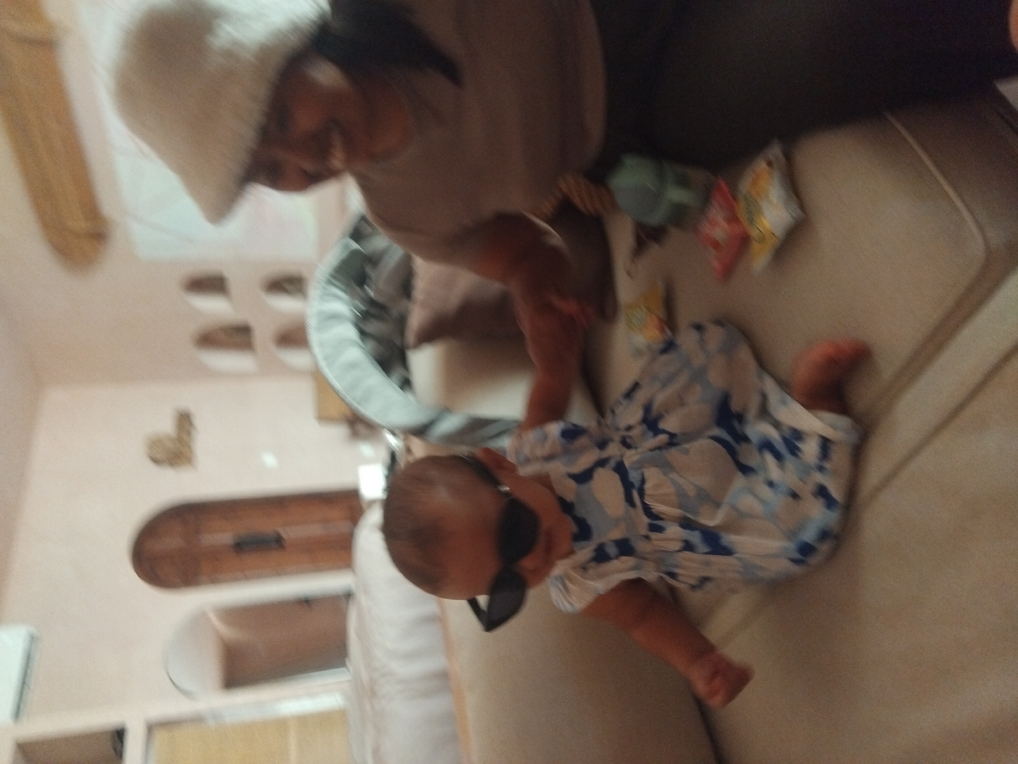 A horizontally rotated indoor shot features an adult and a baby. The adult, wearing a light-colored hat and a beige top, appears to be interacting with the baby. The baby, clad in a white and blue patterned garment and dark sunglasses, is lying down. Several items, including packages and a baby carrier, are on the surface next to the baby. The background includes an arched doorway and an air conditioning unit. The location is Unknown, Unknown.