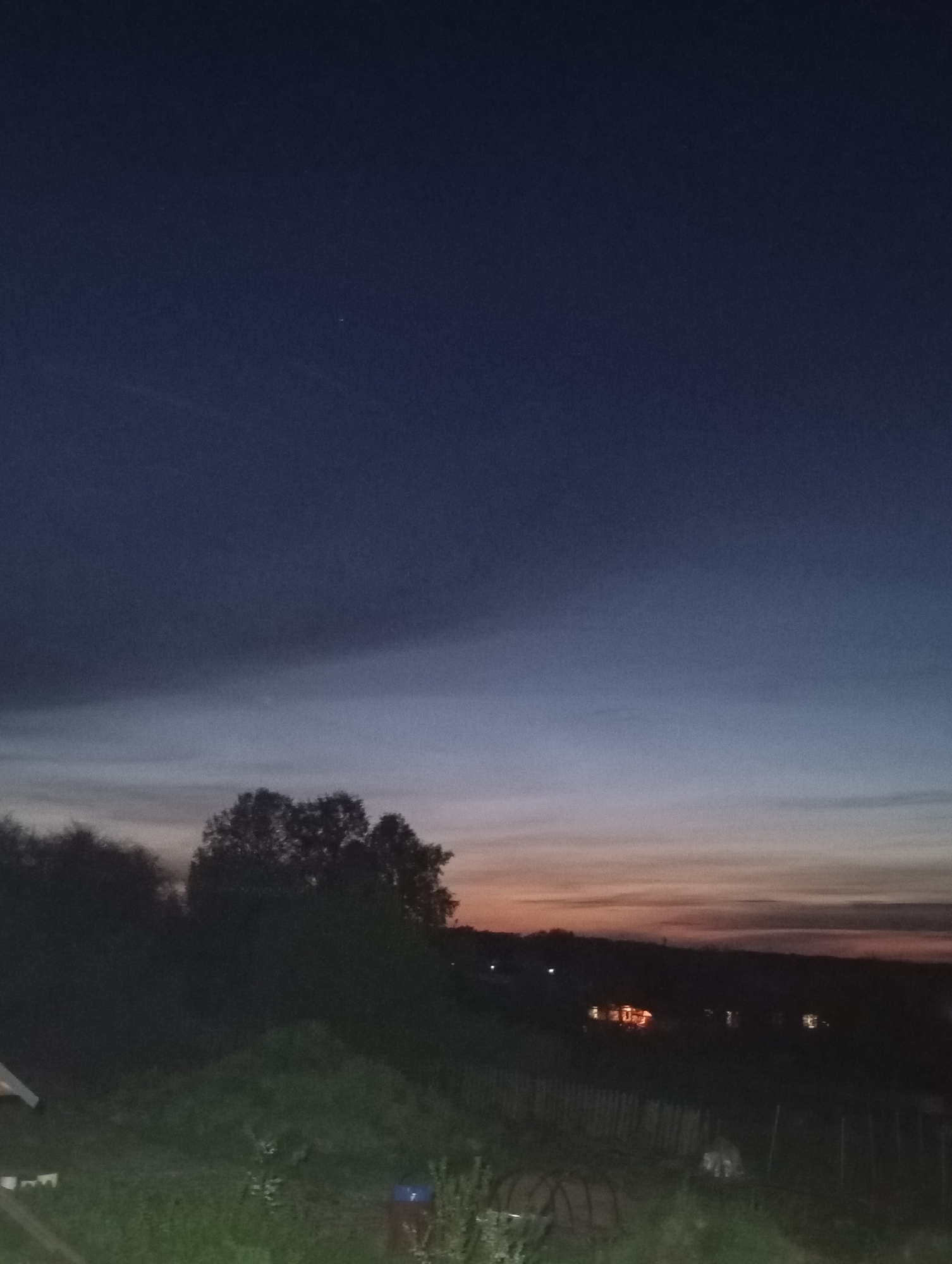 The image shows a rural landscape at dusk or dawn. The sky transitions from a deep blue overhead to a lighter blue and then to an orange horizon. In the foreground, there are trees and vegetation, appearing dark and silhouetted against the sky. Some structures are visible in the mid-ground, including a fence or barrier and what appears to be a small house. The image is underexposed, making details difficult to discern. The location is Unknown, Unknown.