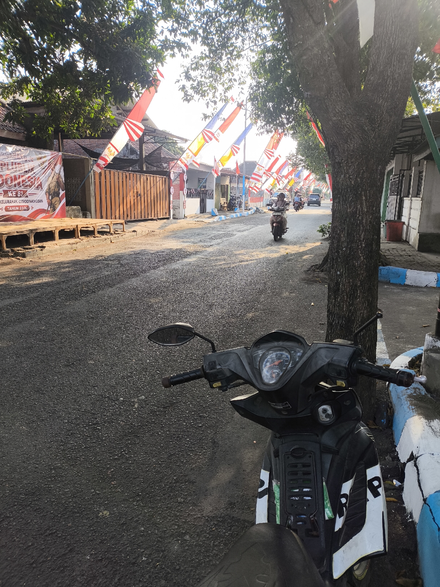 The image depicts a street scene in the city of Citrodiwangsan, Indonesia. In the foreground, a black motorcycle is parked, facing forward. The motorcycle has a detailed dashboard with a speedometer and various indicators. The street is paved with asphalt and lined with a curb painted in alternating blue and white sections.

Along the street, on the left side, there is a large banner with text that appears to be in Indonesian, including "KE 81", "KELURAHAN CITRODIWANGSAN", and "TAHUN 2026". Adjacent to the banner is a wooden structure. Further down the street, buildings are visible, some with wooden gates.

On the right side of the street, a large tree with a rough, textured bark stands prominently. Behind the tree, a building with windows and a red trash bin is situated.

The street is decorated with numerous Indonesian flags, characterized by red and white horizontal stripes. These flags are attached to poles and strung along the street, creating a festive atmosphere.

Several individuals are present on the street, primarily riding motorcycles. One person is visible riding a brown motorcycle further down the road, heading away from the camera. Other motorcycles and at least one car are also present in the mid-ground of the image.

The lighting suggests it is daytime, with sunlight casting shadows across the street, particularly from the tree. The overall environment appears to be a residential or semi-commercial area within the city.