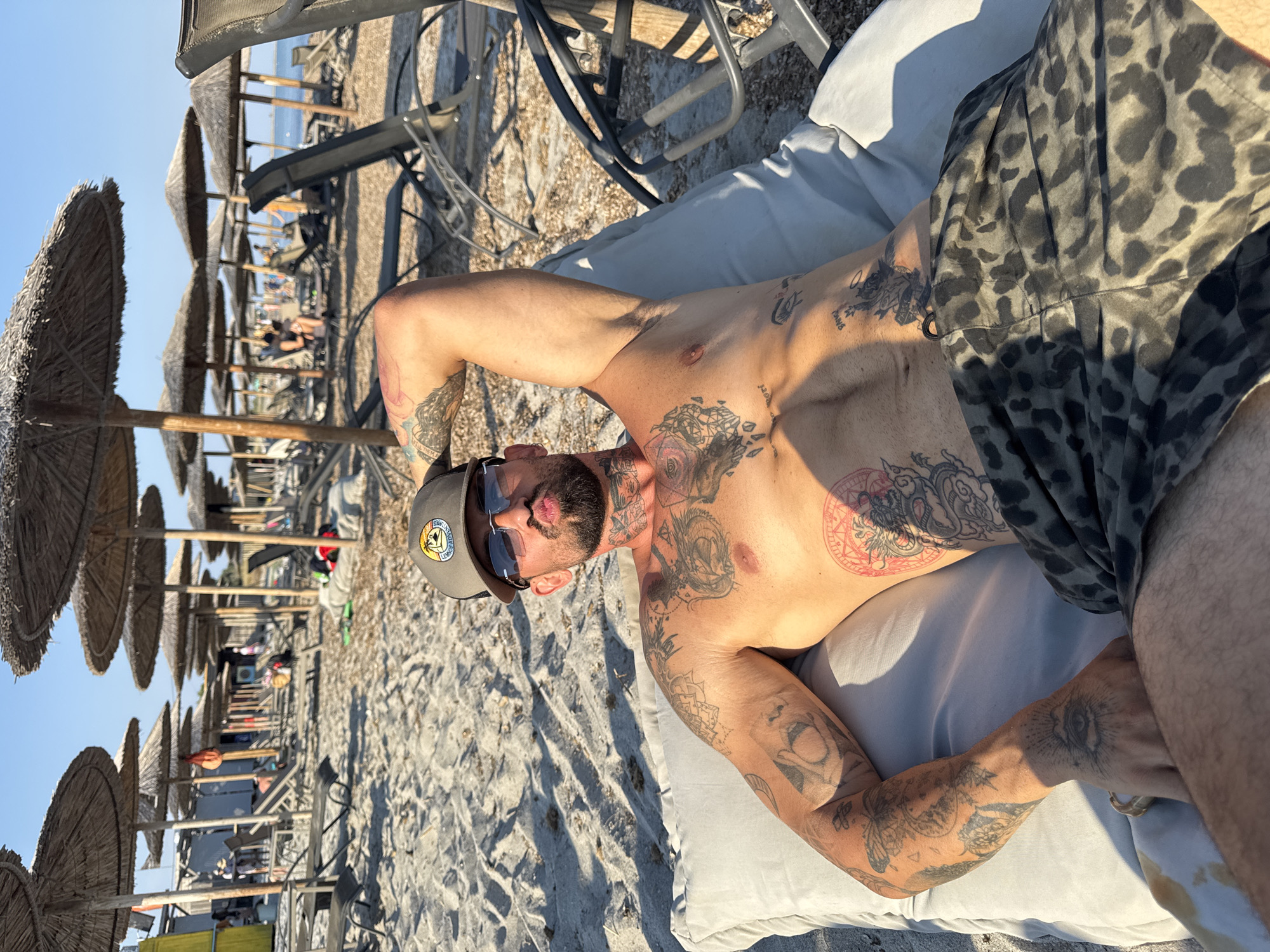 The media file shows a man lying on a cushioned lounger on a sandy beach. He is shirtless and has extensive tattoos covering his chest, arms, and abdomen. He is wearing leopard-print shorts and a baseball cap. Sunglasses are perched on his nose, and he is looking towards the camera. The sun is shining brightly, casting shadows on the sand, indicating it is daytime. In the background, rows of thatched umbrellas and beach chairs suggest a resort or public beach area. The sea and the sky are visible in the distance. The overall impression is one of relaxation and leisure in a sunny, coastal environment. The location context provided is Moudros Municipal Unit, Greece. There is text visible on the man's cap: a circular logo with an image and some writing that is not clearly legible.