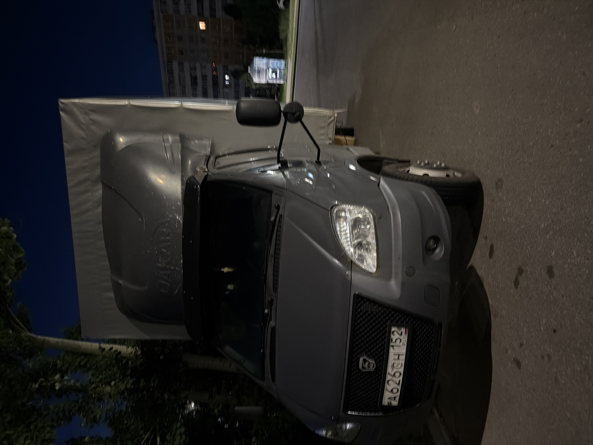 A grey GAZelle Next van is parked on an asphalt road at night. The van has a white tarp covering the cargo area, and a "DAKAR" logo is visible on the front of the cargo cover. The license plate reads "A626CH 152 RUS". The van is positioned at an angle, with its front facing slightly towards the left of the frame. In the background, there is a multi-story residential building with illuminated windows, and trees line the left side of the image. The sky is dark blue, indicating nighttime. There are no people visible in the image.