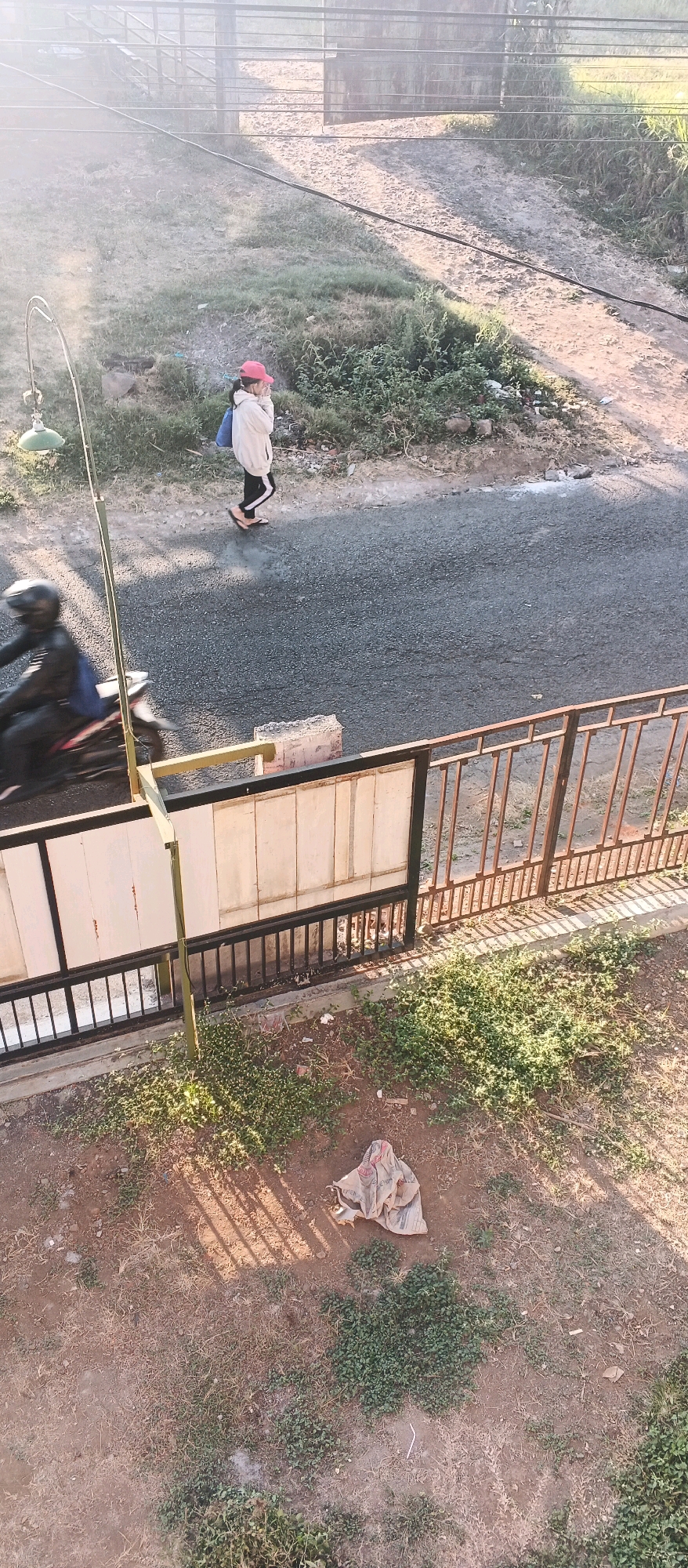 In Bocek, Indonesia, a woman wearing a pink cap, a cream-colored hooded jacket, black pants with white stripes, and flip-flops walks on the side of a paved road. She carries a blue bag over her shoulder and appears to be covering her face with her hands. To her left, a person on a motorcycle, wearing a black helmet and jacket, rides past. A street lamp with a green shade is visible on the left side of the image. In the foreground, a metal fence with vertical bars separates the road from an area with sparse green vegetation and dirt. A piece of beige fabric, possibly a sack, lies on the ground in the foreground, casting a shadow from the fence. The scene is bathed in the warm light of what appears to be early morning or late afternoon.