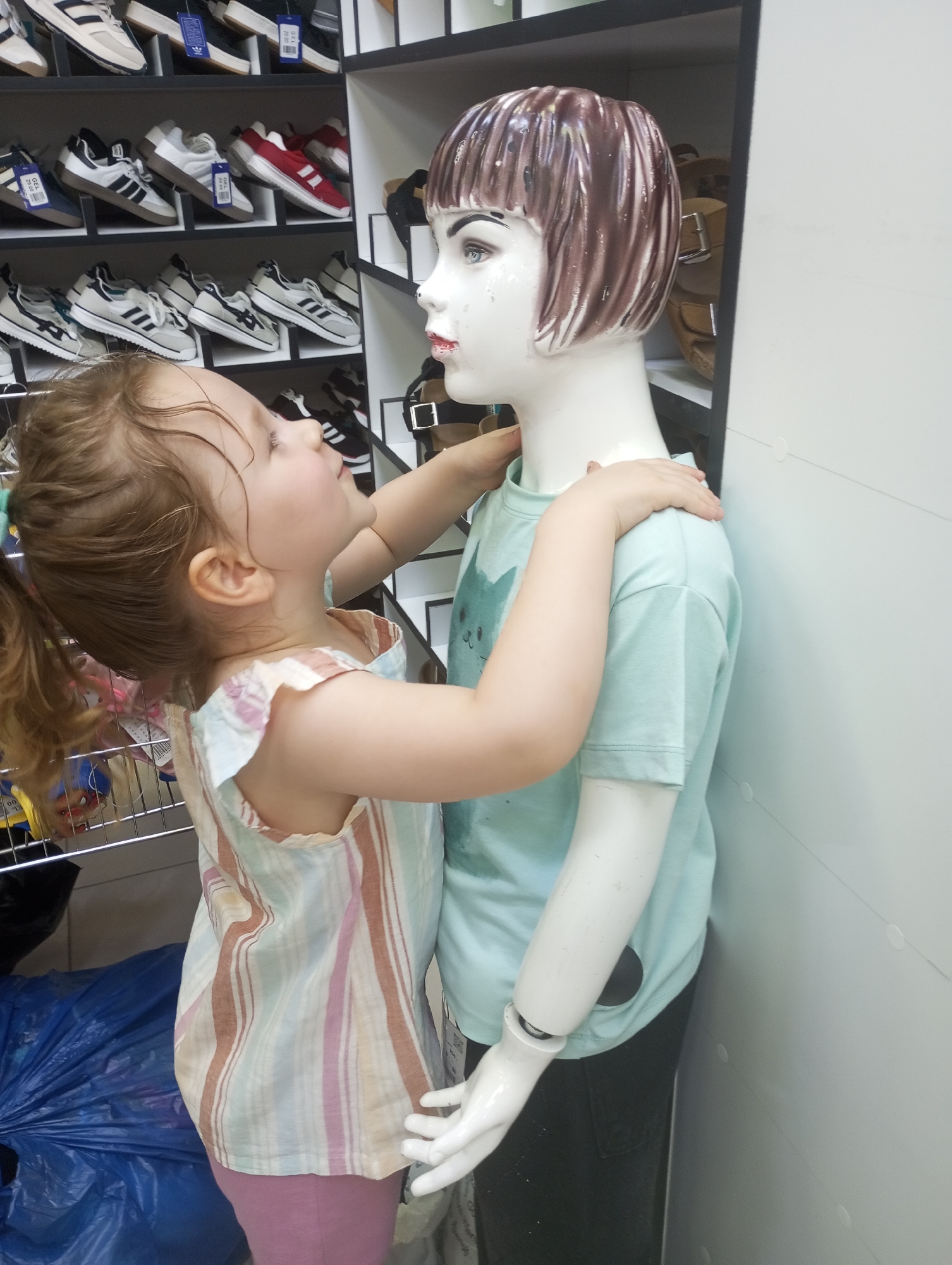 A young girl with blonde hair pulled into a ponytail is interacting with a mannequin in a store. The girl is wearing a striped dress with pink, white, and brown stripes and a pink skirt. She is looking up at the mannequin with a curious expression and has her hands on the mannequin's shoulders. The mannequin is wearing a light blue t-shirt with a cat graphic and black pants. It has short brown hair with lighter streaks.
The background of the image shows shelves filled with shoes, suggesting the setting is a shoe store. The shelves are organized with various styles of sneakers. To the left of the girl, there is a wire rack with some items on it, including what appears to be a blue plastic bag. The overall lighting suggests it is daytime.
The scene captures a playful moment between a child and a display, highlighting the girl's engagement with her surroundings in a retail environment. The location cues, such as the shoe displays, are consistent with a store. There is no visible text in the image that provides specific information about the brand or pricing.