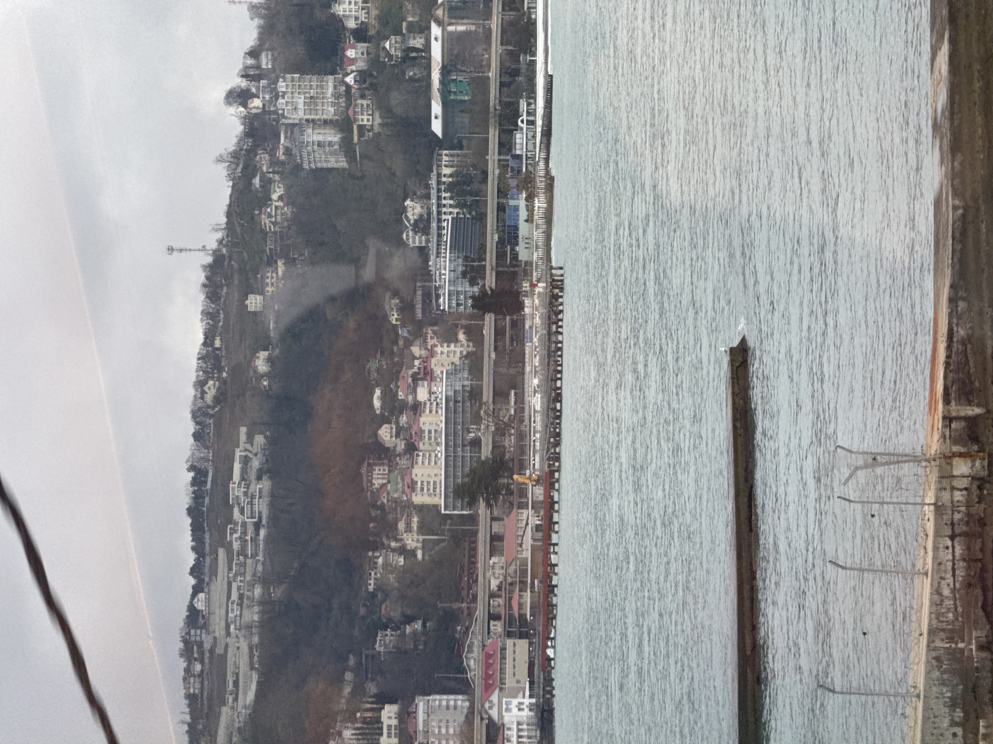 The image shows a coastal view of Sochi, Russia, on a cloudy day. The Black Sea occupies the right side of the frame, its surface textured by slight ripples. A concrete pier or breakwater extends into the sea along the right edge, with metal rungs visible on its side, likely for access. A dark, aged wooden post stands in the water near the pier. The coastline is densely populated with buildings and vegetation climbing up the hillside. The architecture consists of a mix of low-rise and high-rise structures, suggesting a combination of residential and commercial areas. Visible among the buildings are what appear to be hotels, apartment complexes, and possibly some office buildings. A few cranes are visible, implying construction is underway. The atmosphere is hazy. The overall scene is of an urbanized coastal area. A thick, dark cable-like object is visible in the upper-left corner of the image.
