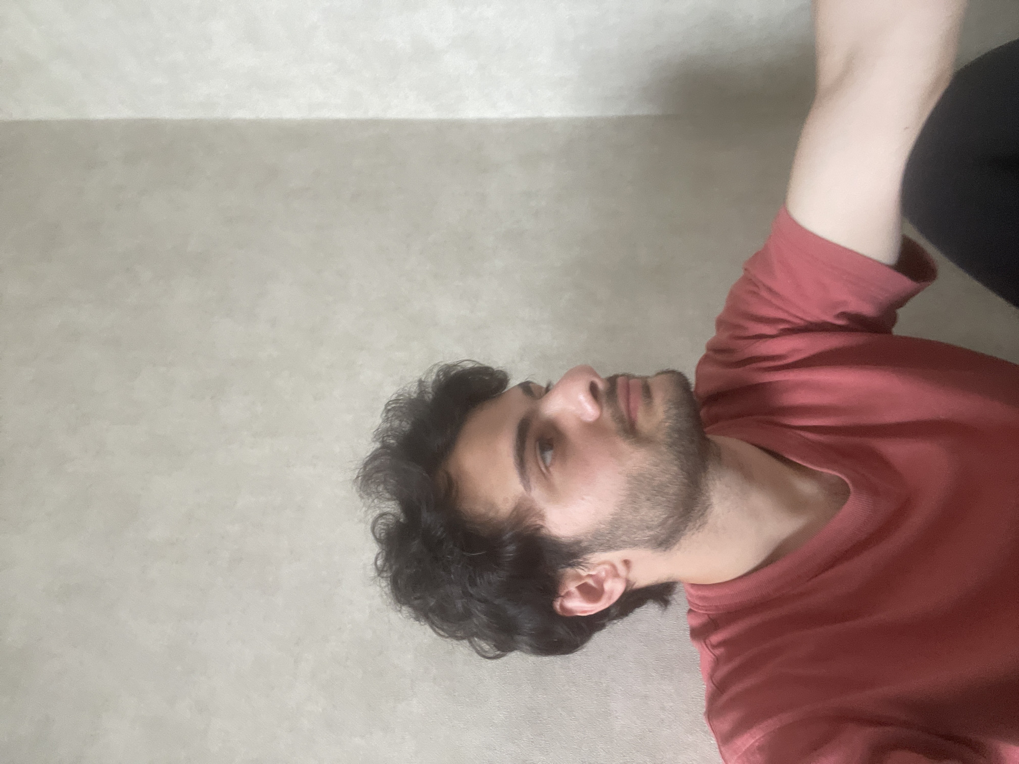 A young man with dark, curly hair and a light beard is pictured from the chest up, facing right. He is wearing a salmon-colored t-shirt. The background is a plain, textured wall that appears to be in a residential setting, possibly an apartment. The lighting is soft and diffused. No specific location in Tbilisi, Georgia, can be identified from the image.