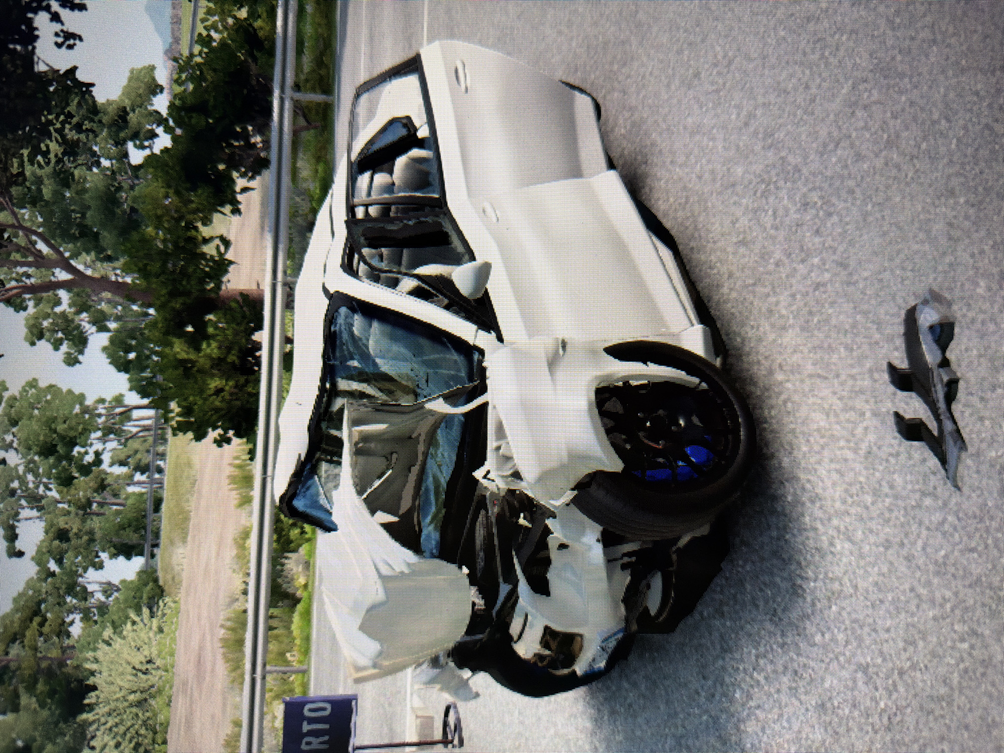 A severely damaged white car is depicted after a crash, lying askew on what appears to be a paved road or track. The image itself appears to be a photograph of a screen, evidenced by the visible pixel grid.
The primary subject is a white car, heavily impacted on its front and driver's side. The entire front end is crushed and deformed, with the hood crumpled and the bumper shattered. The front left wheel is dislodged and angled inward, revealing bright blue brake calipers. The driver's side door and windshield area are also severely compromised, with parts appearing torn or missing. Black debris, likely from the car, is scattered on the asphalt surface to the right of the vehicle.
In the background, a metal guardrail runs along the side of the road, behind which lush green trees and varied vegetation are visible under natural daylight. This suggests an outdoor setting, possibly a rural road or a racing circuit. A small, dark blue sign is partially visible in the lower left corner, with the white letters "RTO" readable. No people are present in the scene. The overall impression is of a static moment immediately following a significant automotive accident.