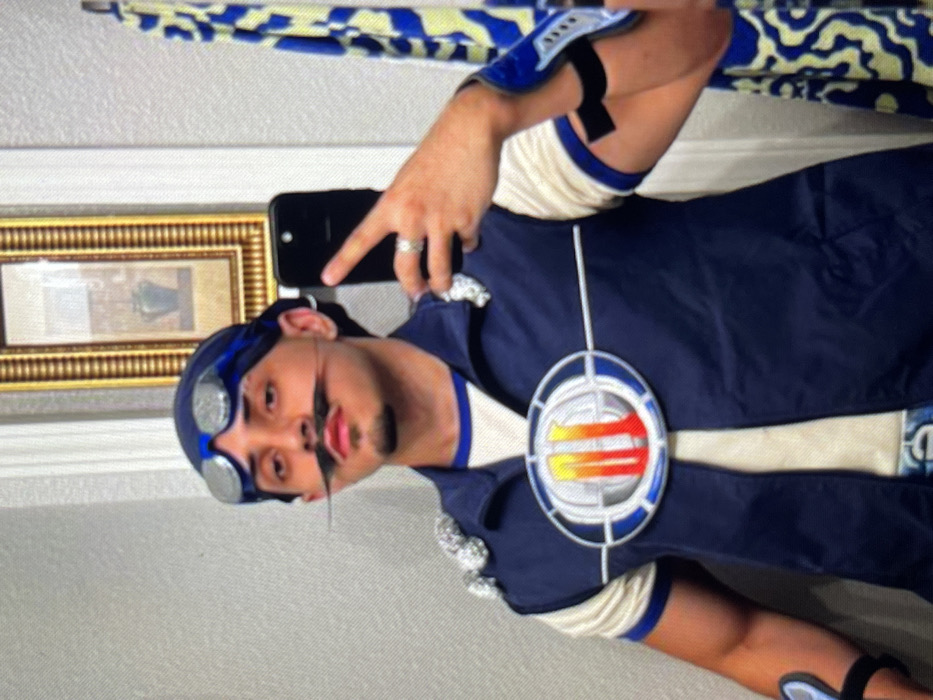 Location: Muchiya, Nigeria.
A male individual is depicted from the chest up, positioned directly in front of a mirror, capturing a selfie with a black smartphone held in his right hand. He wears a blue bandana and silver-framed goggles pushed up onto his forehead. A prominent black handlebar mustache is affixed to his upper lip, accompanied by a small soul patch on his chin. He is dressed in a dark blue sleeveless vest featuring a large, silver and orange circular emblem centered on the chest, over a cream-colored long-sleeve shirt with blue trim on the cuffs. A blue and silver wrist accessory is visible on his right forearm. The background, reflected in the mirror, shows a light gray, textured wall, a gold-framed picture with a dark image inside, and a yellow and blue patterned towel or fabric hanging on the right. His left hand is not visible. A silver ring is present on the ring finger of his right hand.