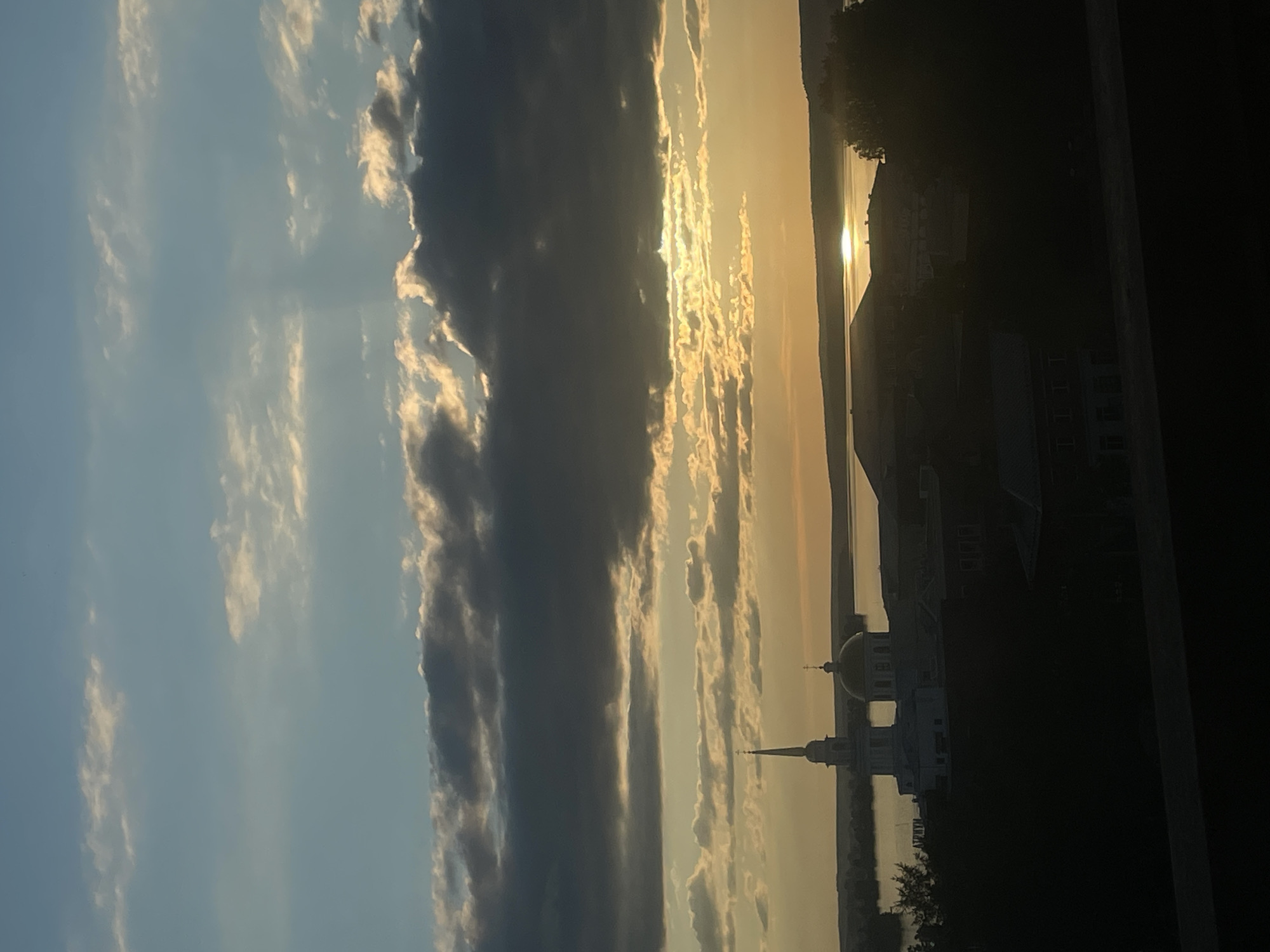 The image, taken in Votkinsk, Russia, captures a serene sunrise or sunset scene over a large body of water, likely the Votkinsk Reservoir. The sky dominates the upper portion of the frame, displaying a gradient of colors from soft blue on the left to warm golden hues near the horizon on the right, indicative of crepuscular light. A prominent band of dark, heavy clouds stretches horizontally across the middle, contrasting with patches of brighter, more illuminated clouds that catch the golden light of the sun. Sunbeams are faintly visible emanating from behind these clouds.
The sun itself is a bright, intense glow, partially obscured by clouds, casting a shimmering reflection across the calm surface of the water. On the far shoreline, silhouetted against the bright horizon, are several buildings and trees. Most notable among these is a white architectural structure featuring a prominent dome and a slender spire, resembling a church or cathedral typical of Russian architecture. This building stands out as a focal point against the warm sky. Other darker, less defined building silhouettes are visible along the distant shore. The overall mood of the image is peaceful and contemplative, highlighting the natural beauty of the landscape at dawn or dusk. There is no visible text in the image.