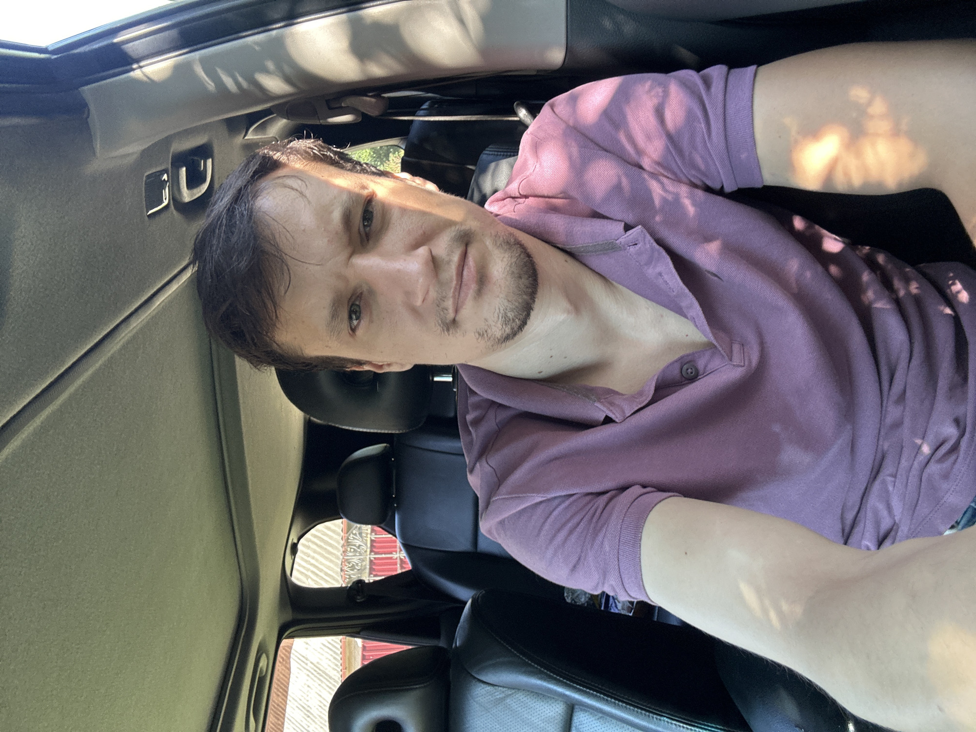 A man with short brown hair and a light beard is sitting in the driver's seat of a car. He is wearing a short-sleeved purple polo shirt. The car interior, including black leather seats and a sunroof, is visible. Sunlight casts dappled shadows on the man's face, arm, and shirt. Outside the car, a building with a red roof and white siding is partially visible. The scene appears to be set in the city of Shatava, Ukraine.